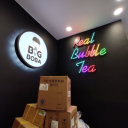 Wymarzony Dom ArtEmi - Czarna ściana z neonowym napisem 'Real Bubble Tea' i okrągłym podświetlanym logo 'Big Boba', poniżej stos kartonów z napisem '700 注塑饮料杯 9070)' i sylwetką kubka, w tle widoczna osoba na drabinie.