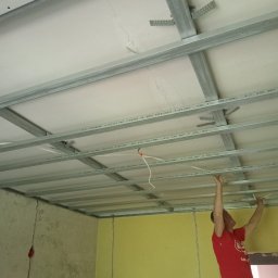Wymarzony Dom ArtEmi - Montaż metalowej konstrukcji sufitu podwieszanego z widocznymi przewodami elektrycznymi, wykonywany przez mężczyznę w czerwonej koszulce, na tle ścian w różnych kolorach i z widocznymi pęknięciami.