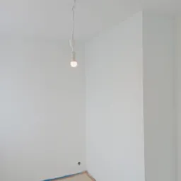 Pomalowane na biało ściany w rogu pokoju, zabezpieczona podłoga, wisząca żarówka bez klosza.