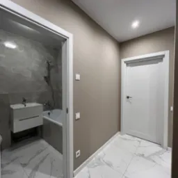 Wykończona łazienka z białymi płytkami imitującymi marmur na podłodze i ścianach, widoczna umywalka wisząca z szafką, wanna, białe drzwi wewnętrzne, beżowe ściany i oświetlenie punktowe.