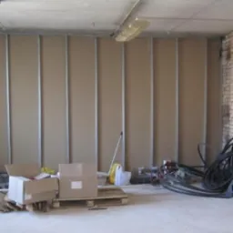 Wnętrze w trakcie prac wykończeniowych, widoczne płyty na stelażu tworzące ścianę działową, kartony na palecie, zwisające kable i narzędzia w nieładzie.