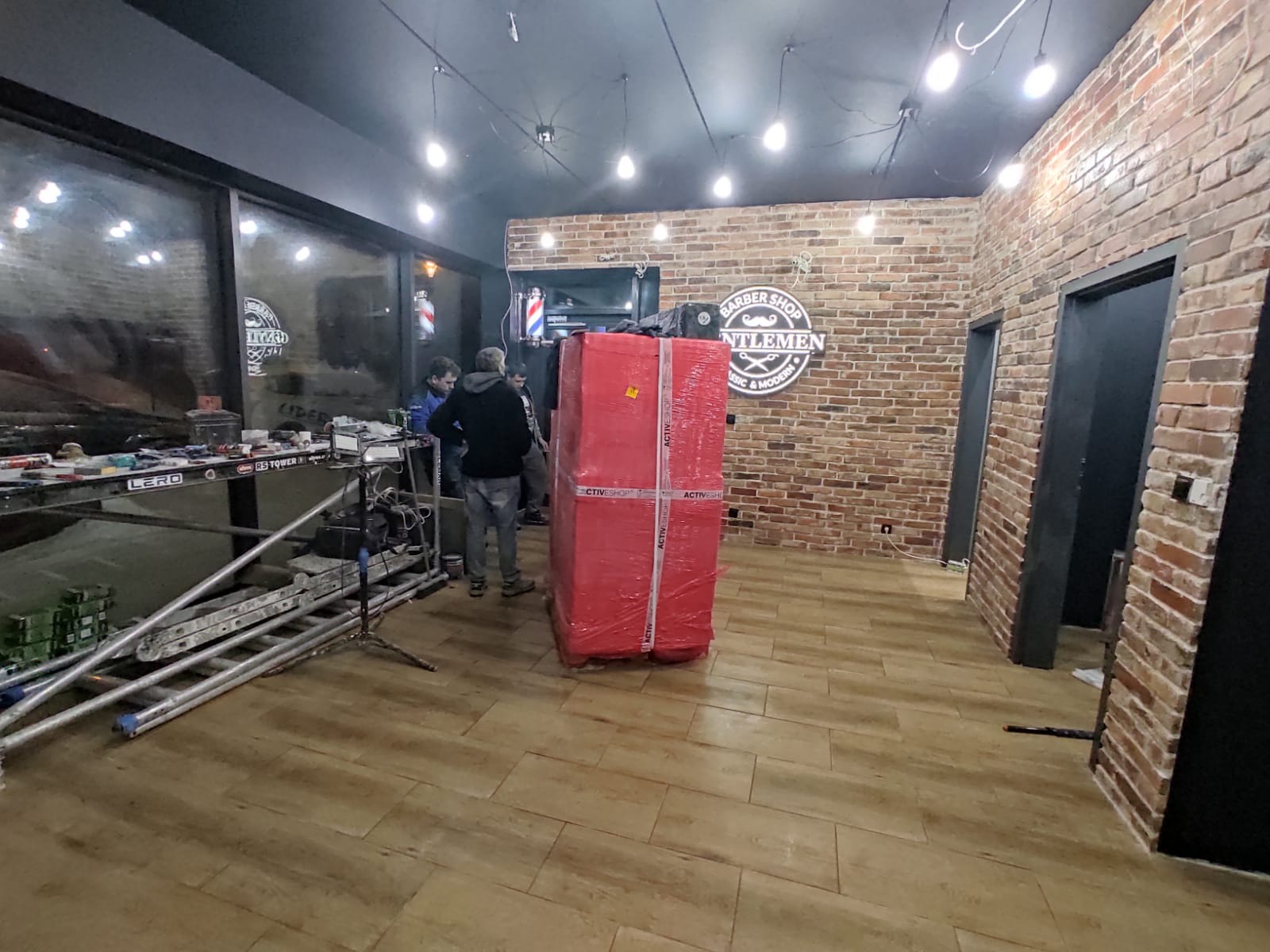 Wnętrze lokalu usługowego w Gliwicach w trakcie prac wykończeniowych, widoczna ceglana ściana, podłoga imitująca drewno, duża paczka owinięta folią, rusztowanie i grupa osób.
