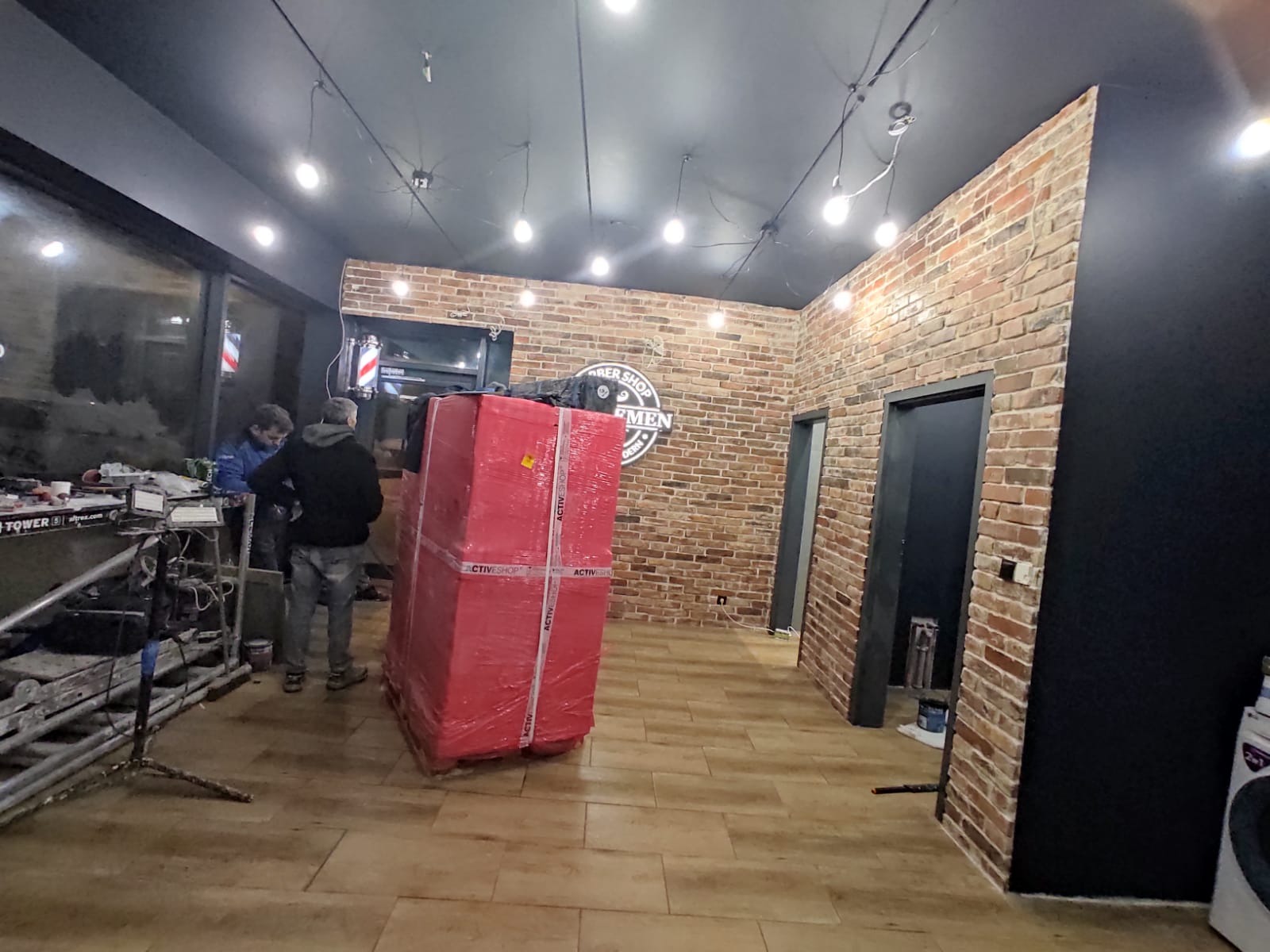 Wnętrze lokalu usługowego w trakcie wykańczania: widoczna ceglana ściana, czarny sufit z oświetleniem punktowym, podłoga imitująca drewno, pracownicy oraz duży, zapakowany w folię mebel...
