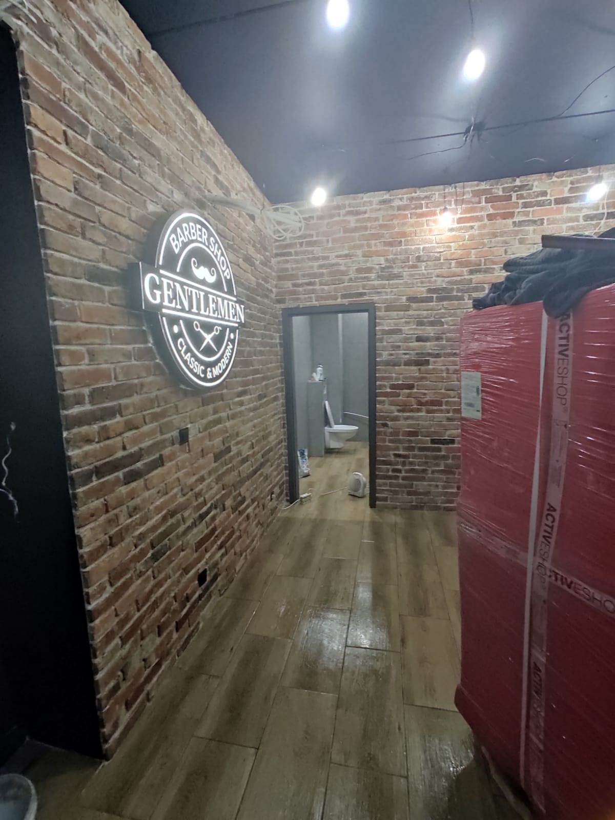 Wnętrze lokalu usługowego z ceglanymi ścianami i podświetlanym logo 'Barber Shop Gentlemen Classic & Modern'. W głębi widoczne drzwi do łazienki z białą toaletą oraz zapakowany towar w folii.