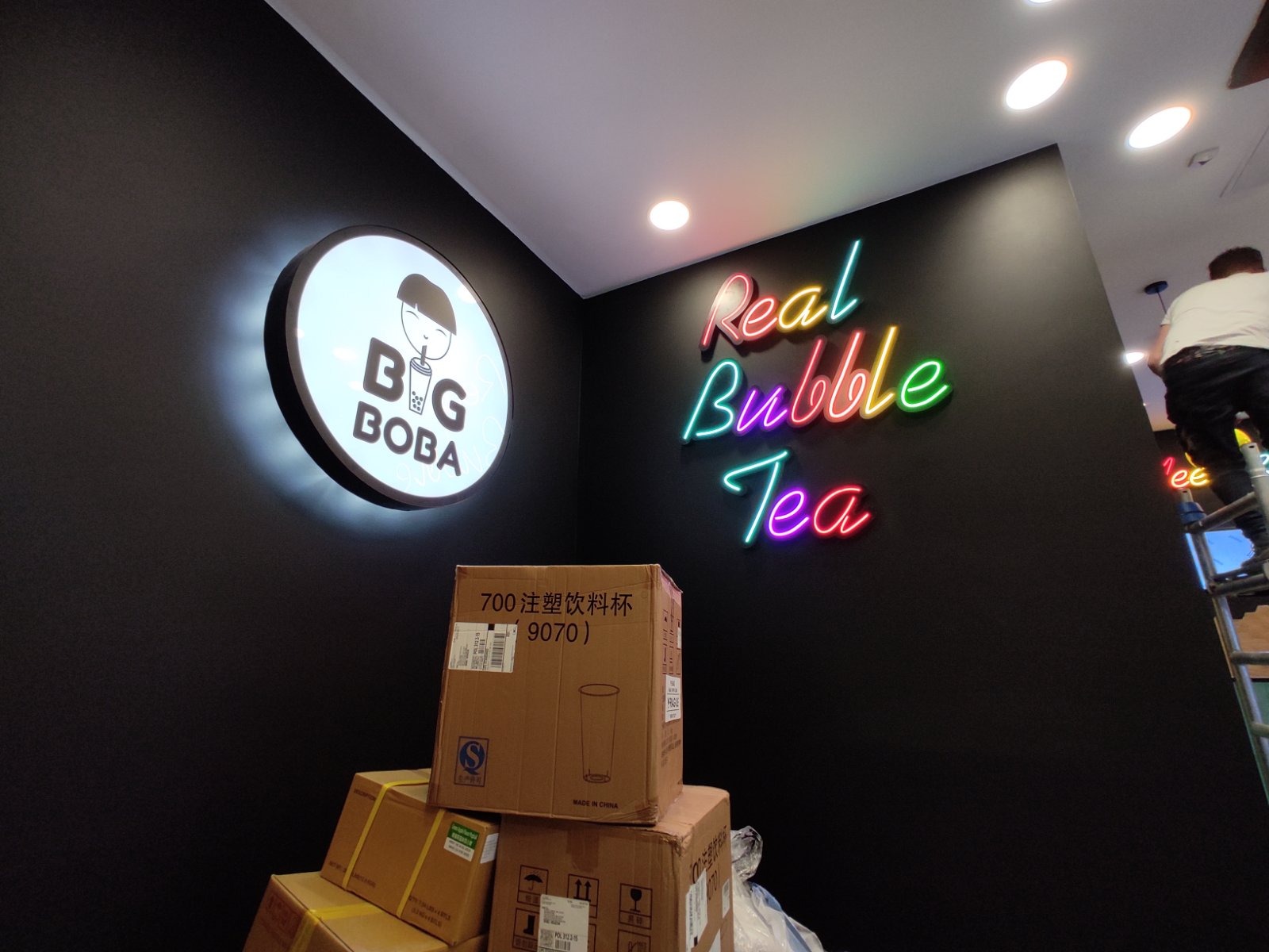 Czarna ściana z neonowym napisem 'Real Bubble Tea' i okrągłym podświetlanym logo 'Big Boba', poniżej stos kartonów z napisem '700 注塑饮料杯 9070)' i sylwetką kubka, w tle widoczna osoba na drabinie.