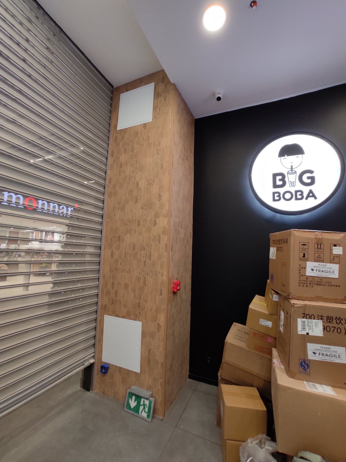 Wykończenie ściany w lokalu z okładziną przypominającą plaster miodu, obok metalowa roleta, logo 'Big Boba' na czarnej ścianie i stos kartonów z napisem 'Fragile'.