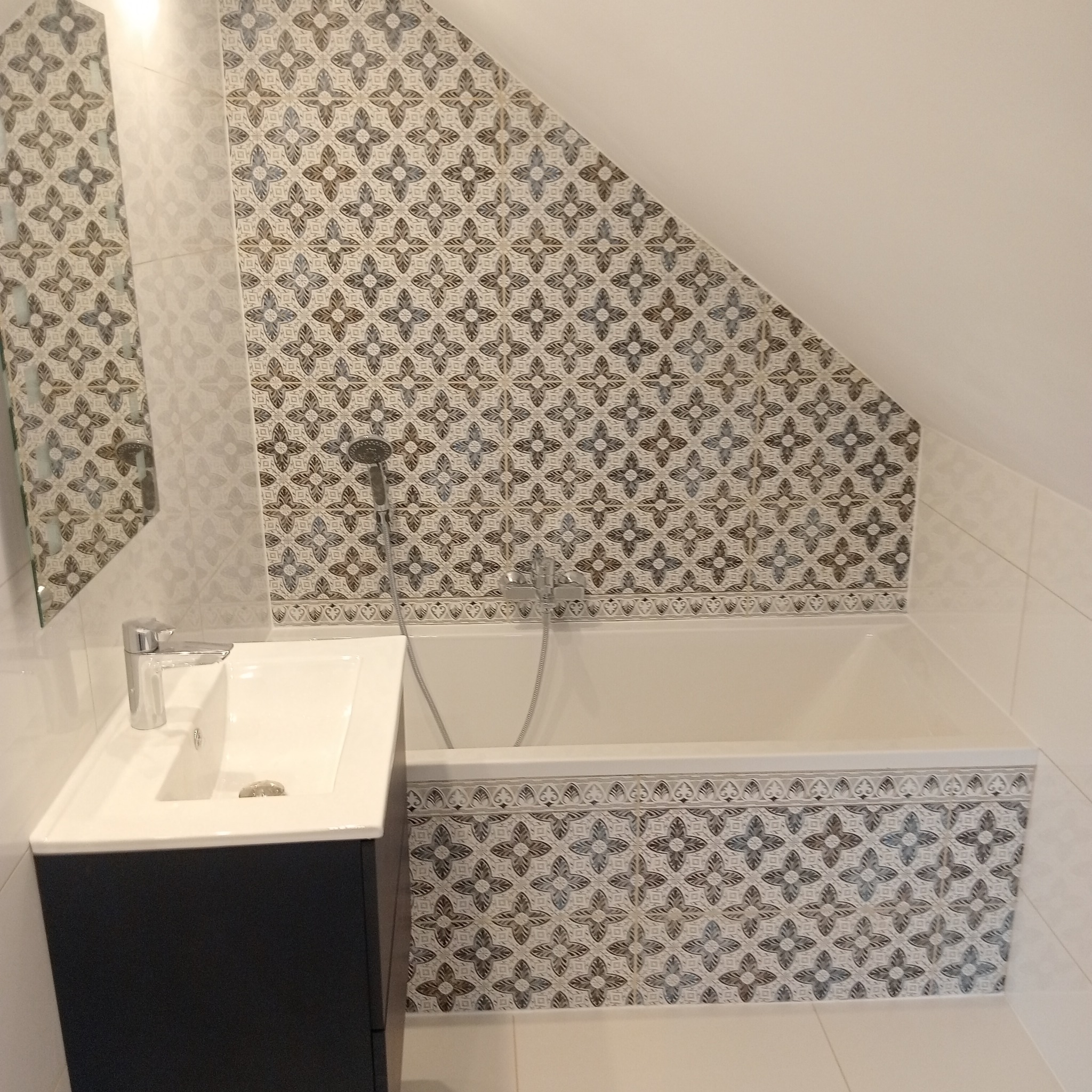 Nowoczesna łazienka na poddaszu z białą wanną obudowaną płytkami w kwiatowy wzór, umywalką z szafką w kolorze antracytowym i chromowaną armaturą.