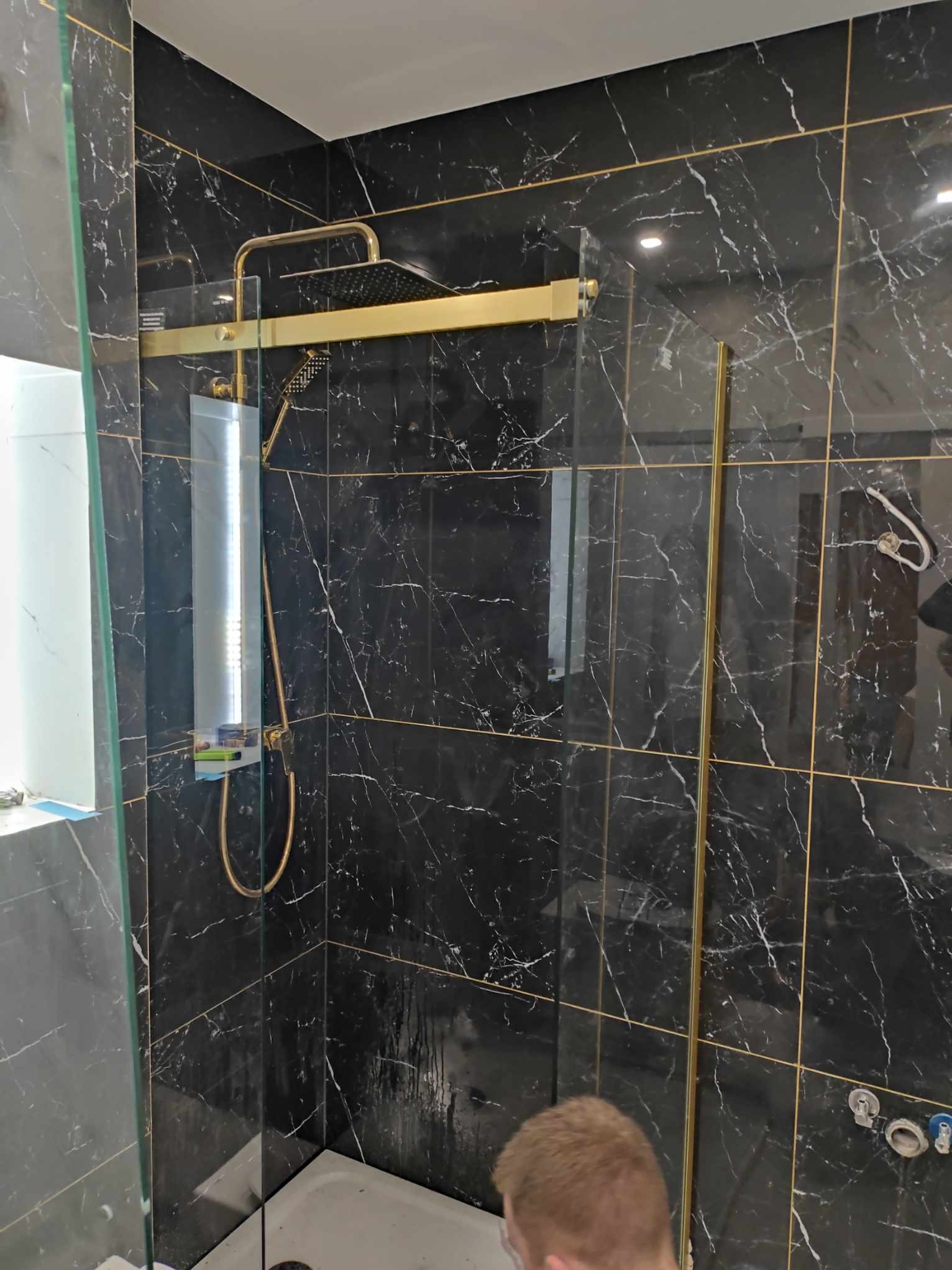 Nowoczesna kabina prysznicowa z czarnymi płytkami imitującymi marmur i złotymi akcentami, widoczna szklana ściana oraz złota armatura.