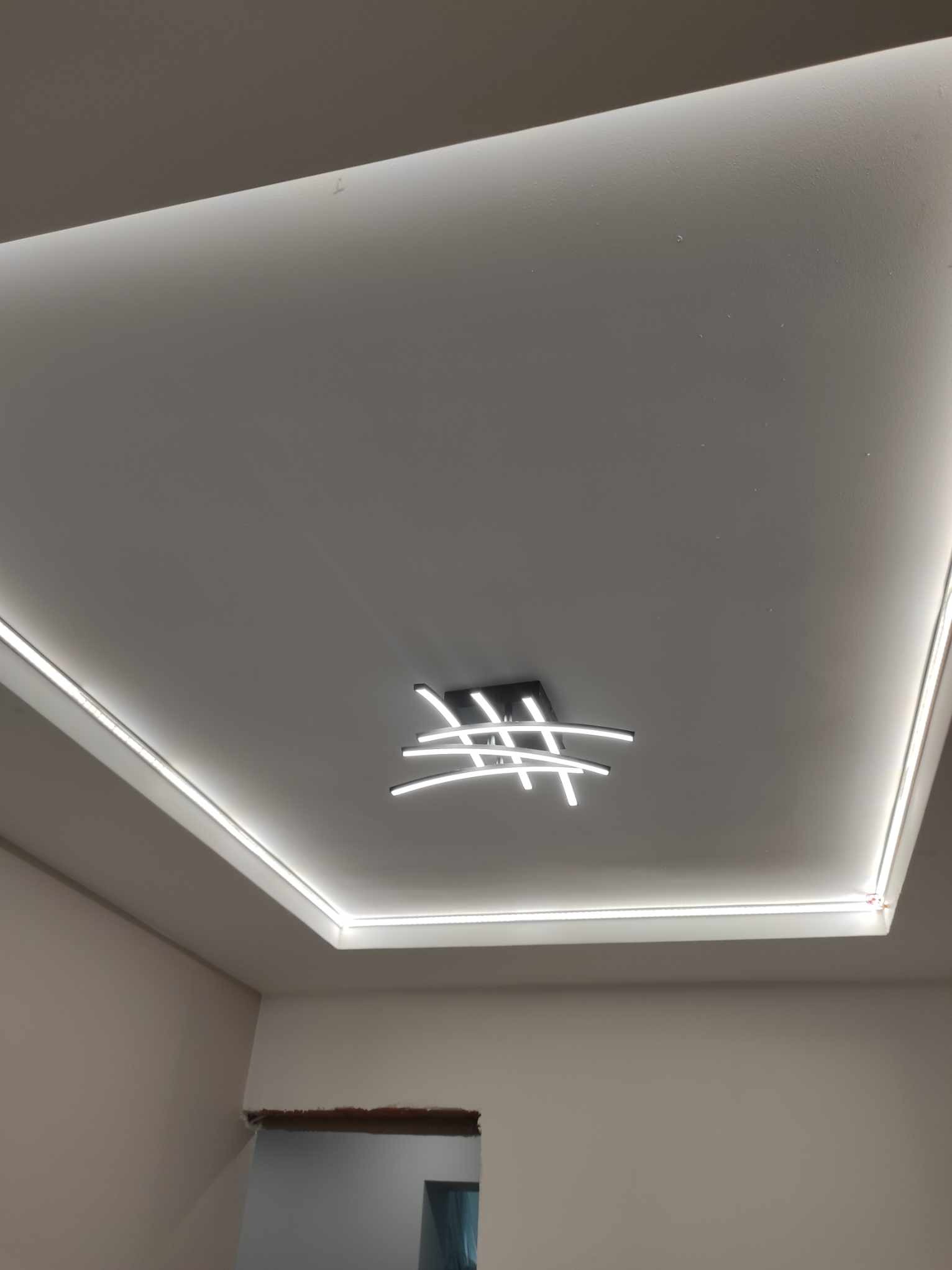 Nowoczesny sufit podwieszany z oświetleniem LED wzdłuż krawędzi i centralnym, designerskim żyrandolem z białych linii.