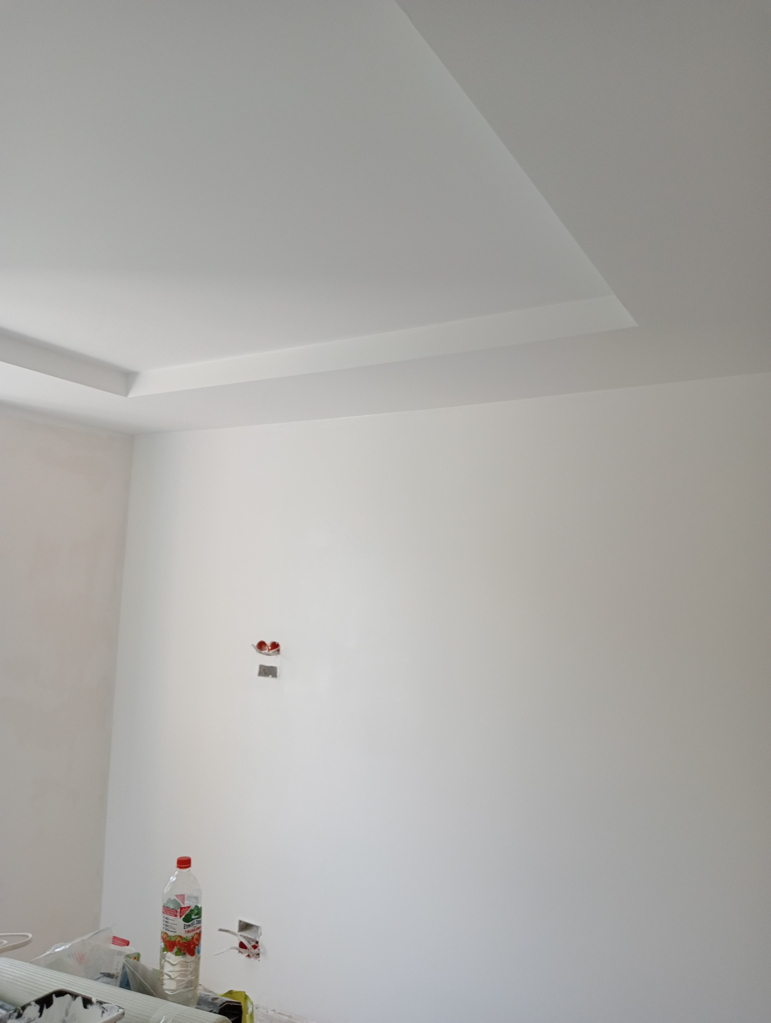 Pomieszczenie w trakcie prac wykończeniowych, widoczne gładkie, białe ściany i sufit z elementami dekoracyjnymi, niezabezpieczone gniazdka elektryczne oraz butelka z napojem i materiały budowlane...
