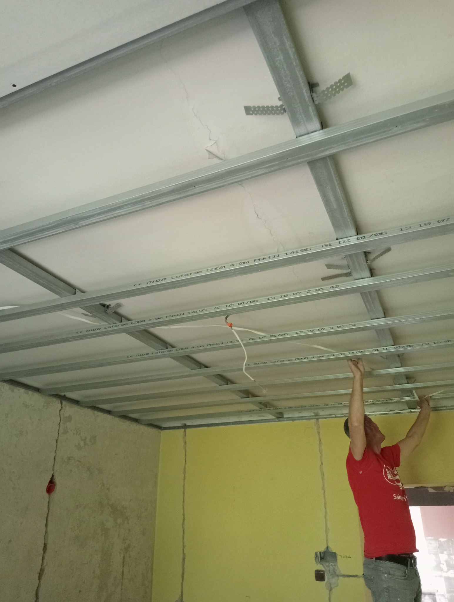 Montaż metalowej konstrukcji sufitu podwieszanego z widocznymi przewodami elektrycznymi, wykonywany przez mężczyznę w czerwonej koszulce, na tle ścian w różnych kolorach i z widocznymi pęknięciami.