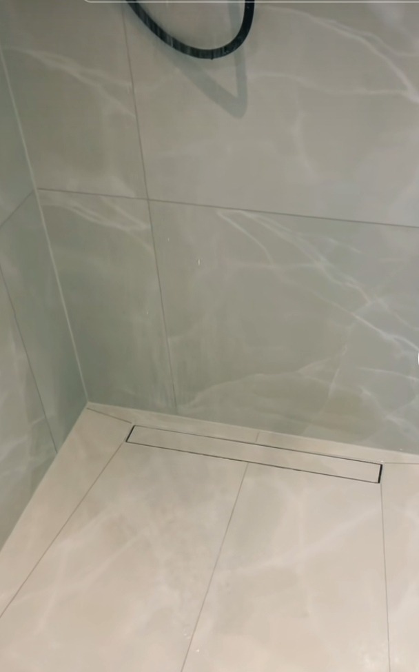 Ukończona podłoga w kabinie prysznicowej z jasnych, marmurkowych płytek ceramicznych i liniowym odpływem podłogowym.