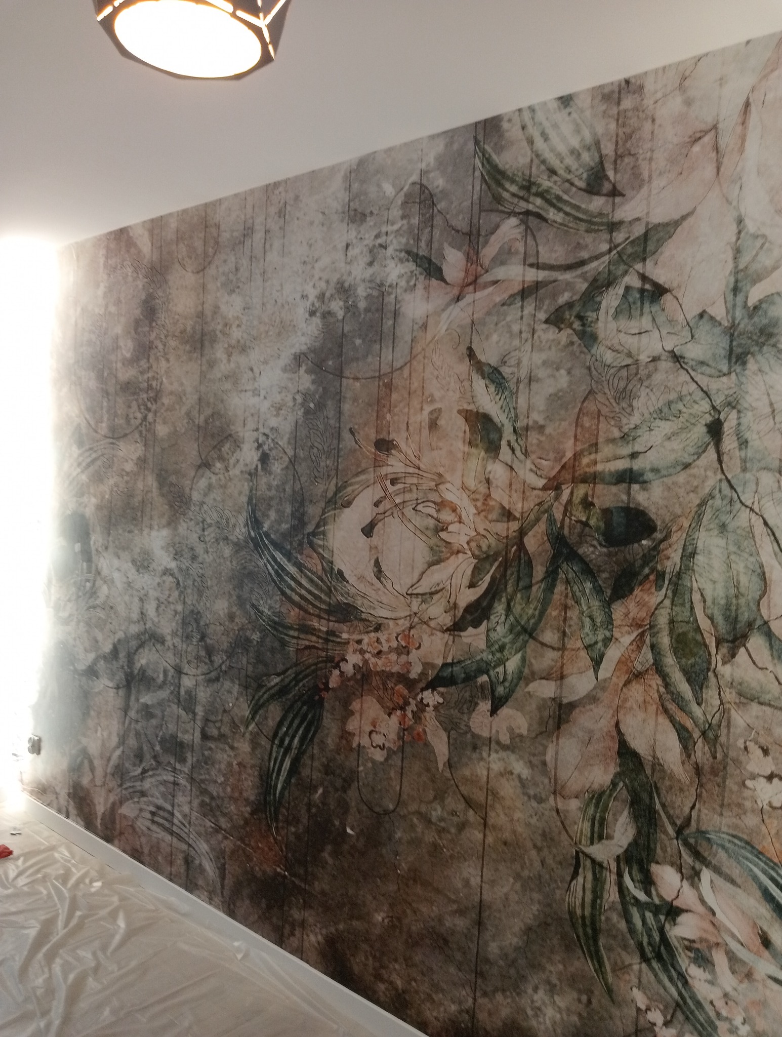 Ściana z fototapetą o motywie kwiatowym w odcieniach brązu i zieleni, zabezpieczona folią podłoga, widoczne gniazdka elektryczne.