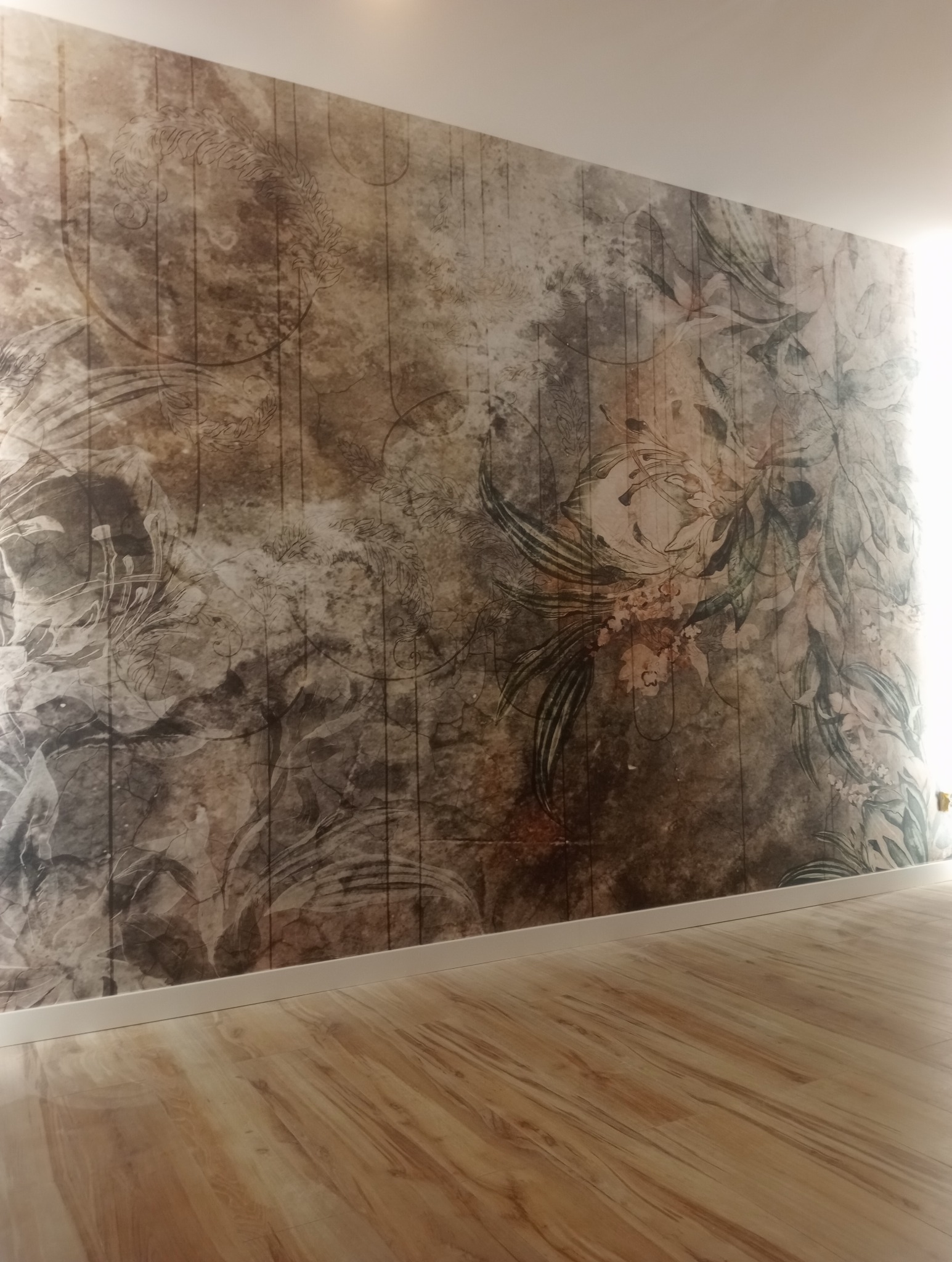 Ściana z fototapetą o wzorze kwiatowym w odcieniach brązu i szarości, z widoczną drewnianą podłogą i białą listwą przypodłogową.