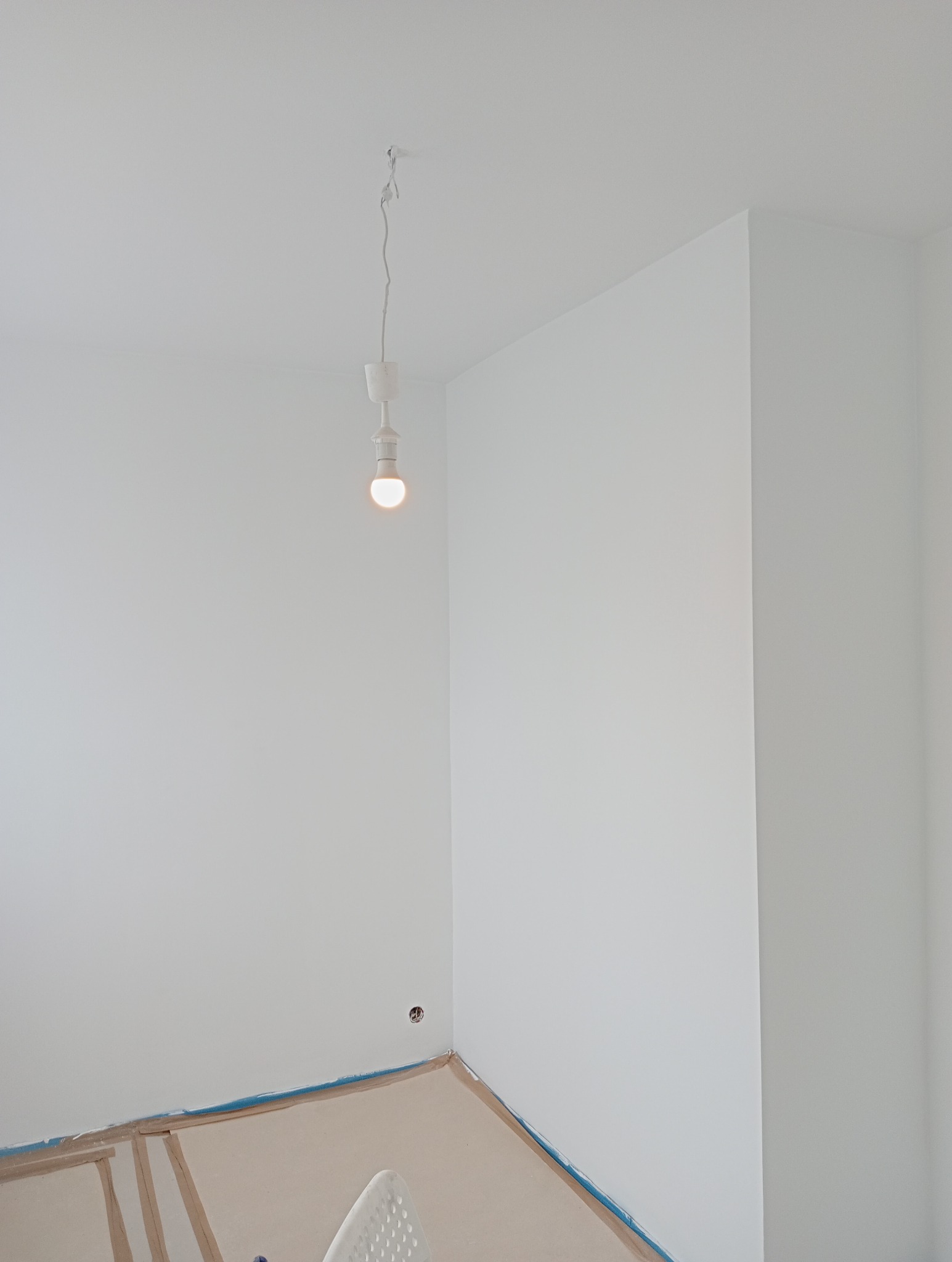 Pomalowane na biało ściany w rogu pokoju, zabezpieczona podłoga, wisząca żarówka bez klosza.