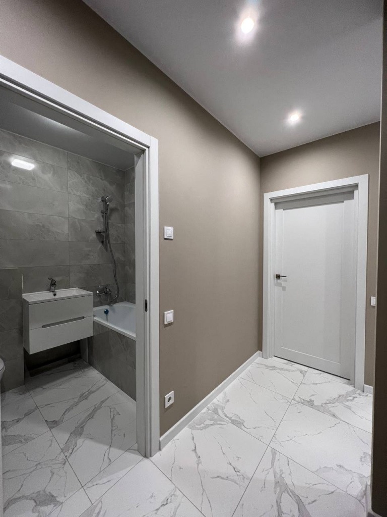 Wykończona łazienka z białymi płytkami imitującymi marmur na podłodze i ścianach, widoczna umywalka wisząca z szafką, wanna, białe drzwi wewnętrzne, beżowe ściany i oświetlenie punktowe.