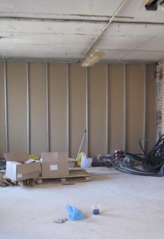 Wnętrze w trakcie prac wykończeniowych, widoczne płyty na stelażu tworzące ścianę działową, kartony na palecie, zwisające kable i narzędzia w nieładzie.