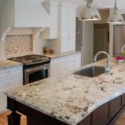 Kamieniarstwo GRANBUD Waldemar Rycyk - Kuchenna wyspa z jasnym, nakrapianym blatem, chromowanym zlewem i baterią, otoczona białymi szafkami z kryształowymi gałkami i widoczną kuchenką gazową.