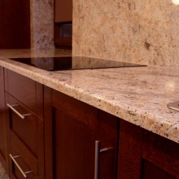 Kamieniarstwo GRANBUD Waldemar Rycyk - Jasny, nakrapiany blat z kamienia w kuchni z ciemnymi, drewnianymi szafkami i wbudowaną płytą grzewczą.