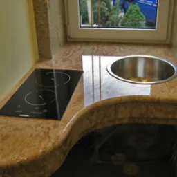 Kuchenny blat kamienny z wbudowaną płytą indukcyjną i okrągłym zlewem ze stali nierdzewnej, widok z góry na jasnym, naturalnym kamieniu z widocznym oknem w tle.