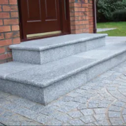 Dwa stopnie wejściowe wykonane z szarego kamienia, z zaokrąglonymi krawędziami, prowadzące do brązowych drzwi wejściowych w ceglanym budynku, z kostką brukową ułożoną w wachlarz na podłożu.