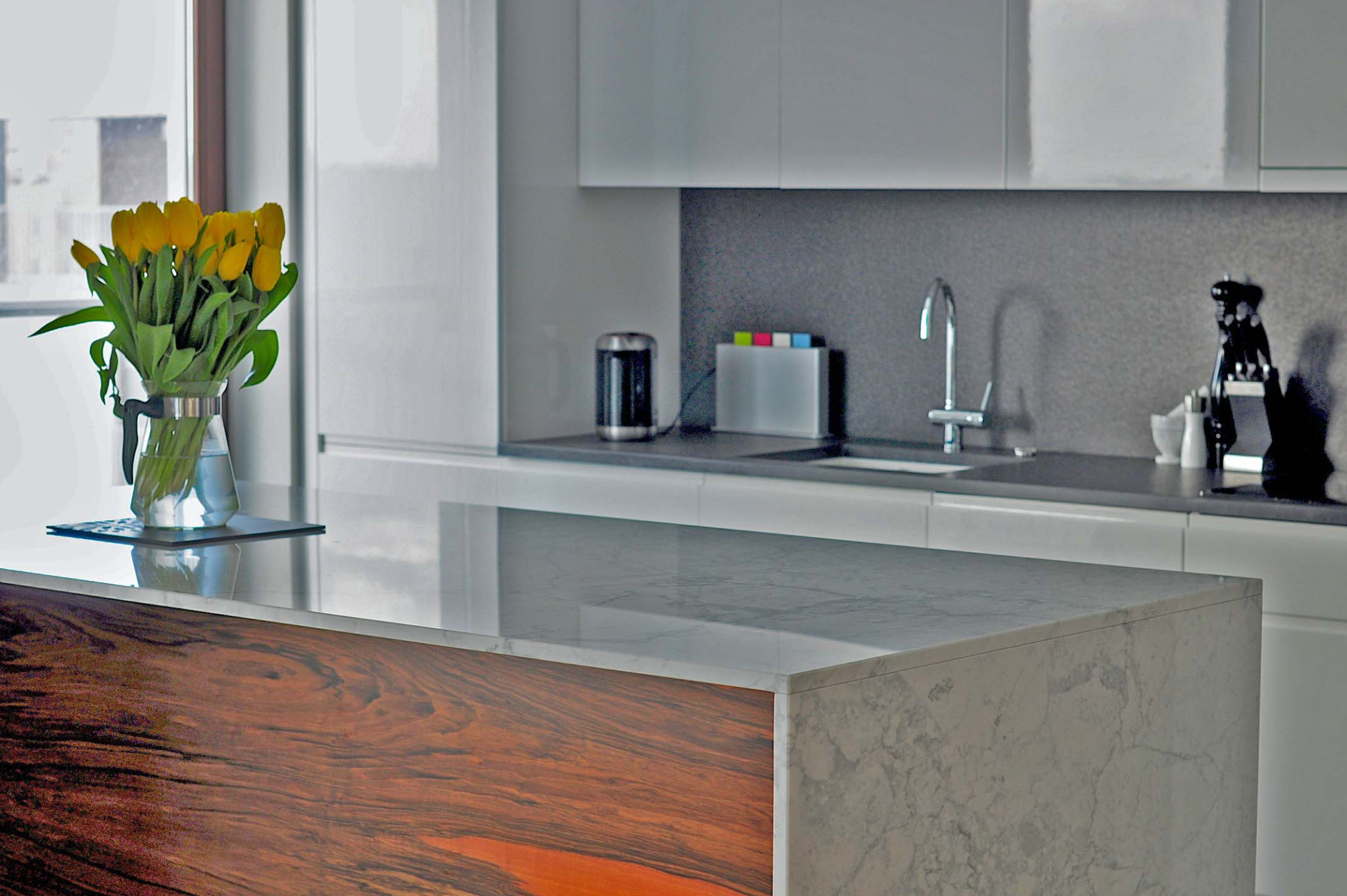 Wyspa kuchenna z blatem z szarego kamienia z widocznym użyleniem, połączona z drewnianą zabudową w odcieniach brązu i pomarańczy, na blacie wazon z żółtymi tulipanami, w tle nowoczesna kuchnia...