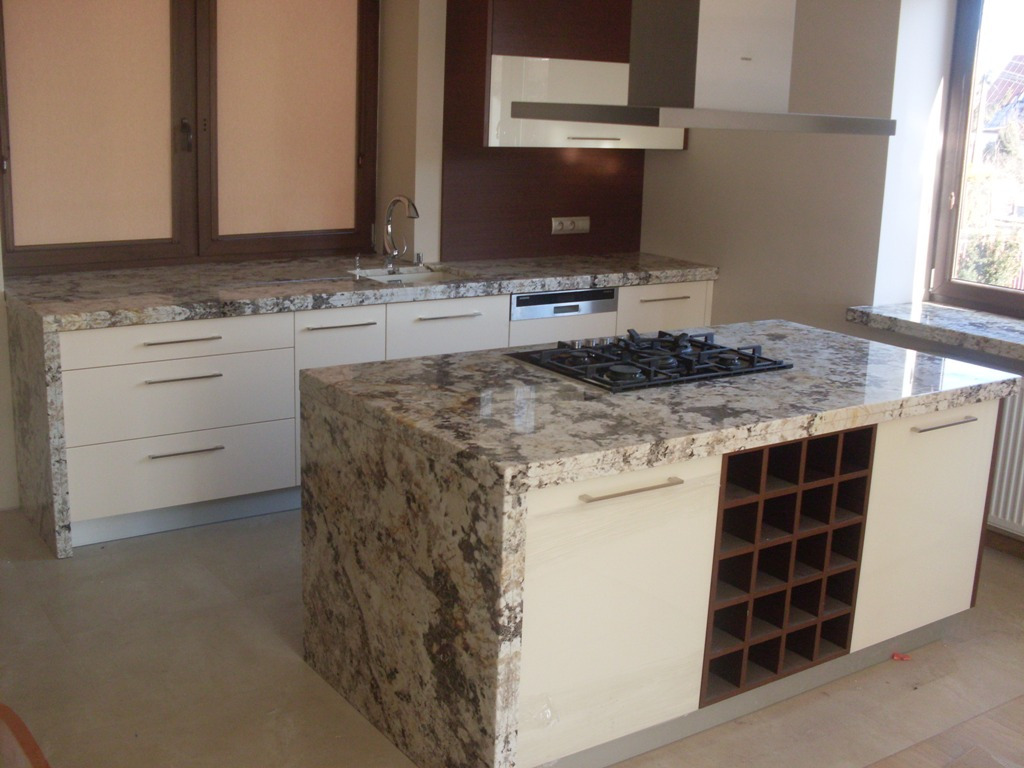 Nowoczesna kuchnia z wyspą, blaty z kamienia o wyraźnym wzorze, wbudowana płyta gazowa, białe szafki z podłużnymi uchwytami, drewniany regał na wino w wyspie, okno z parapetem z tego samego...