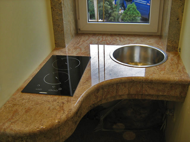 Kuchenny blat kamienny z wbudowaną płytą indukcyjną i okrągłym zlewem ze stali nierdzewnej, widok z góry na jasnym, naturalnym kamieniu z widocznym oknem w tle.