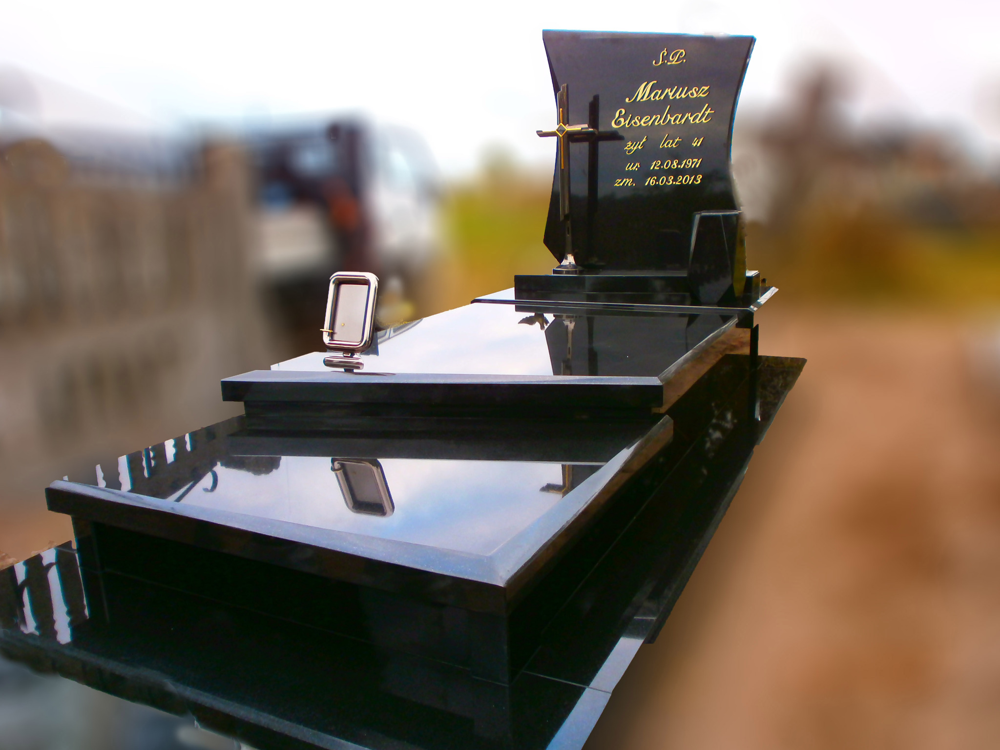 Czarny, polerowany pomnik z wygrawerowanym złotym napisem 'Mariusz Eisenbardt' i datami urodzenia i śmierci, zwieńczony złotym krzyżem; mała ramka na zdjęcia po lewej stronie.
