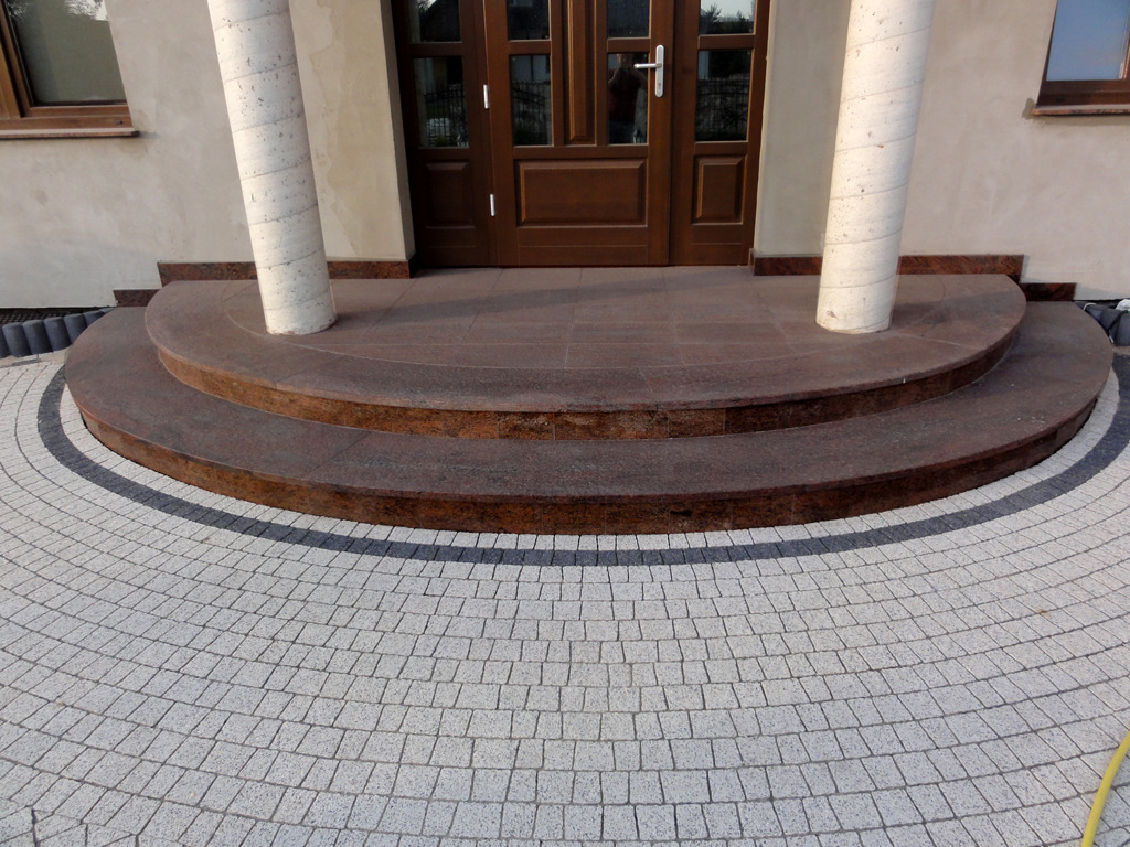 Dwu-stopniowe wejście do domu wyłożone brązowym kamieniem, z okrągłym brukowanym chodnikiem i dwoma kolumnami po bokach brązowych drzwi wejściowych.