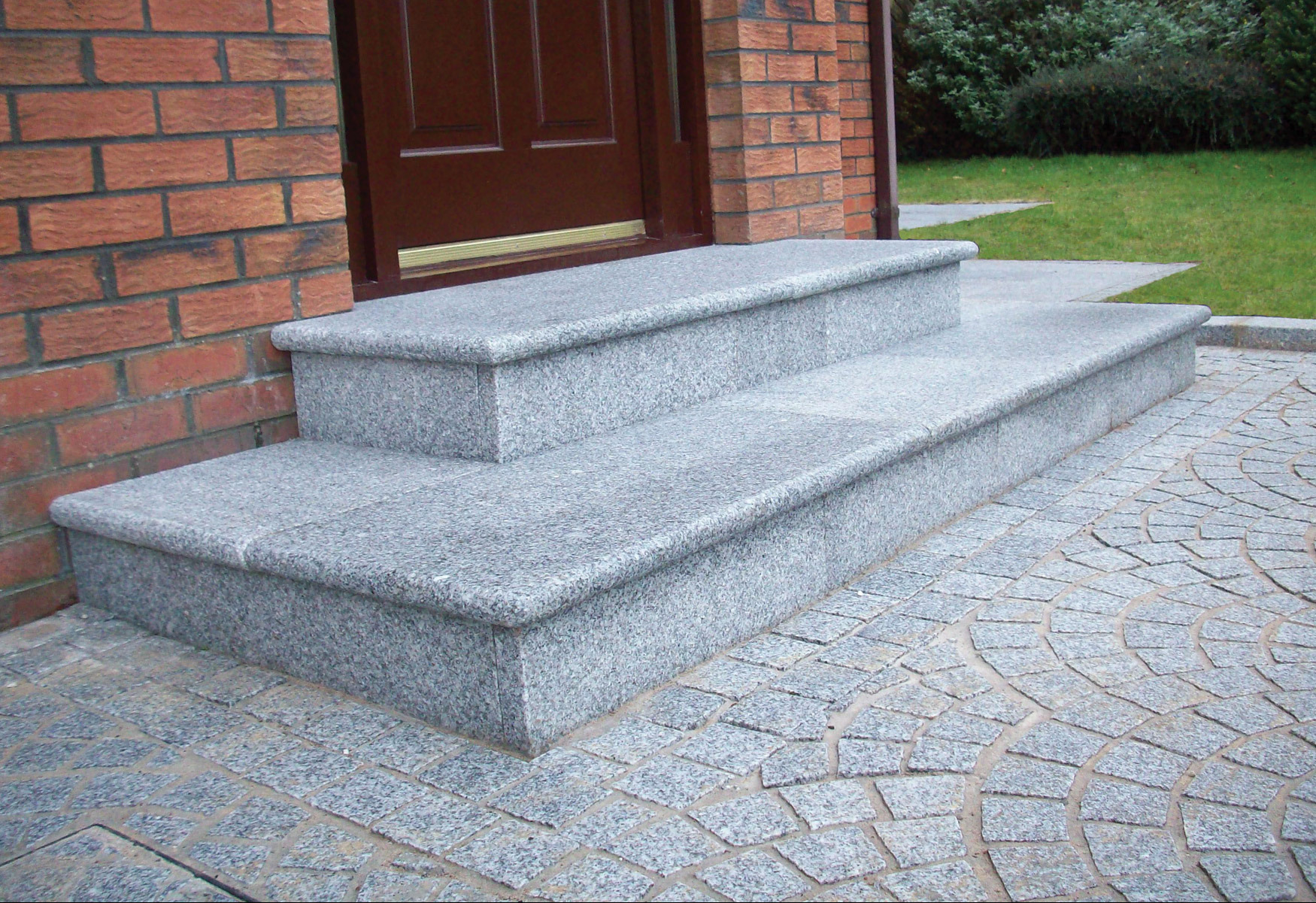 Dwa stopnie wejściowe wykonane z szarego kamienia, z zaokrąglonymi krawędziami, prowadzące do brązowych drzwi wejściowych w ceglanym budynku, z kostką brukową ułożoną w wachlarz na podłożu.