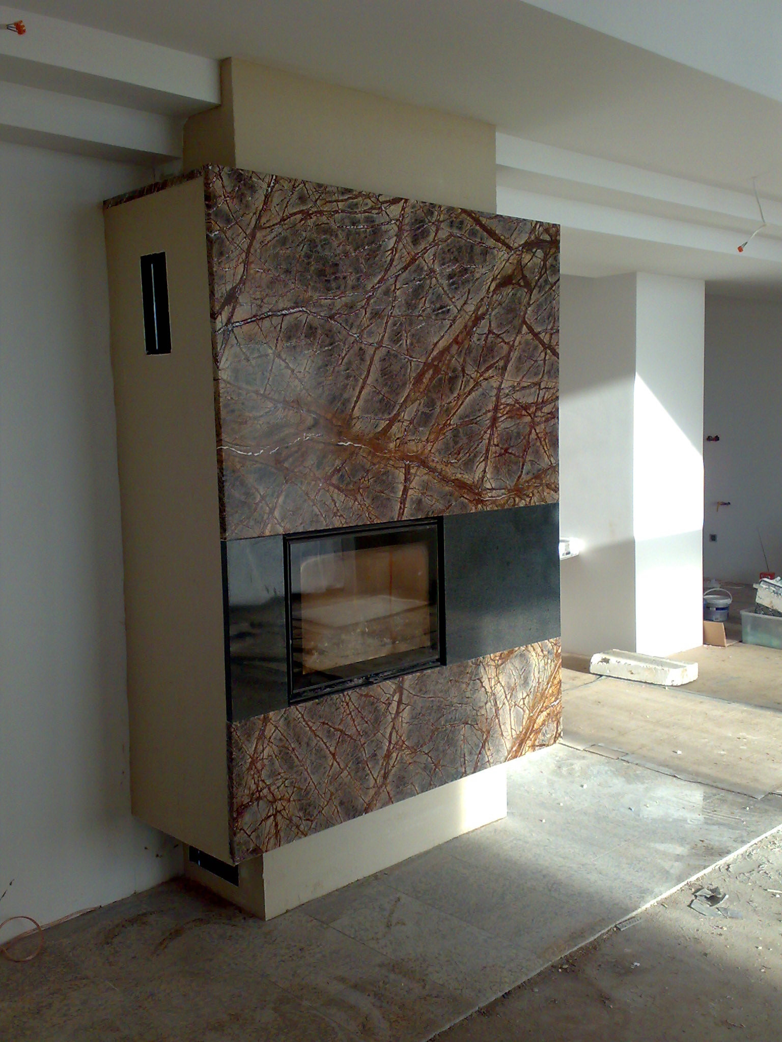 Nowoczesny, obudowany w brązowy marmur z wyraźnym żyłkowaniem, kominek z czarnym wkładem, w surowym wnętrzu w trakcie prac wykończeniowych.
