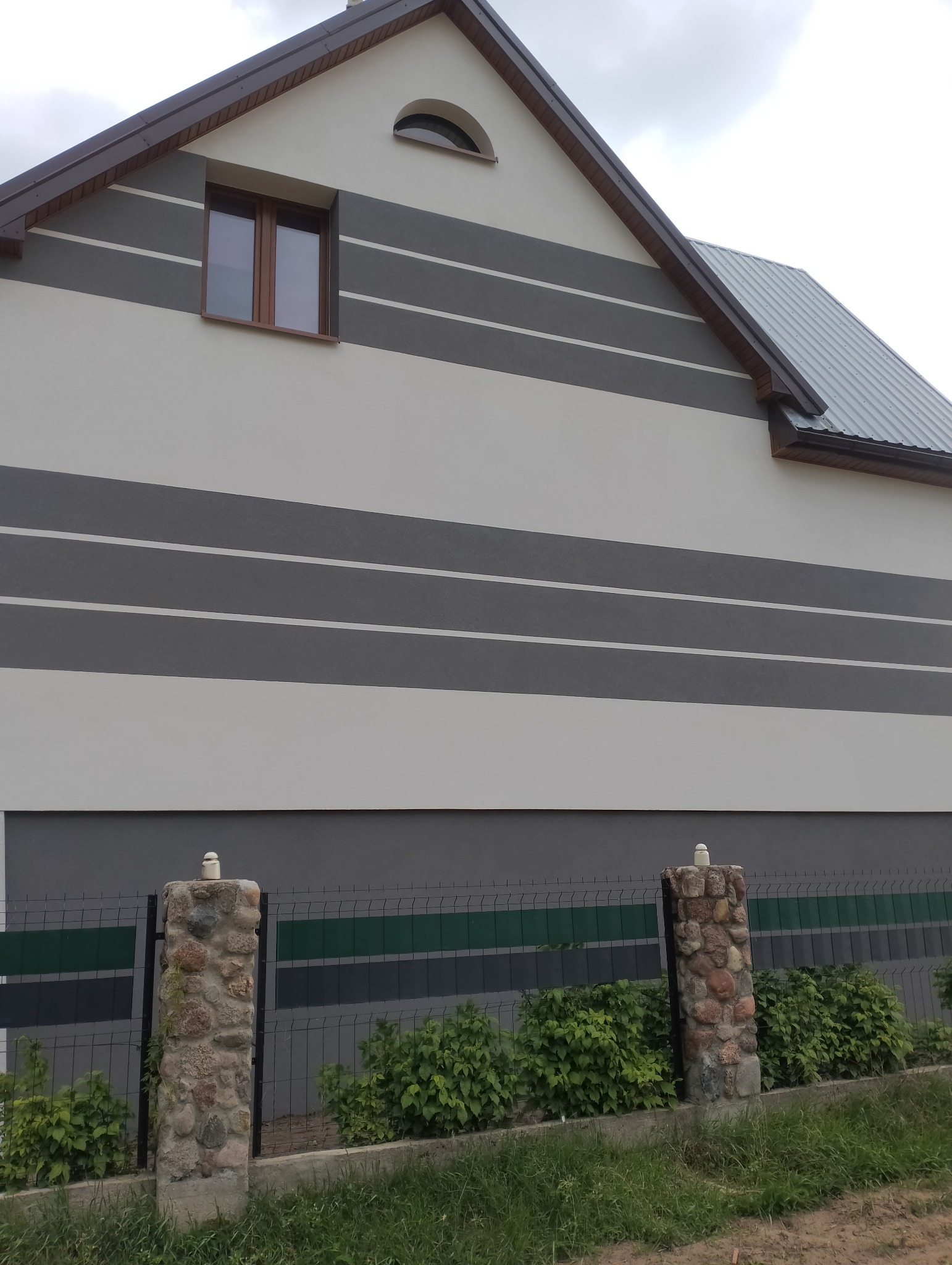 Elewacja domu z szarymi pasami i oknem z brązową ramą, widok z dołu. Kamienne słupki ogrodzenia z zielonymi krzewami na pierwszym planie.
