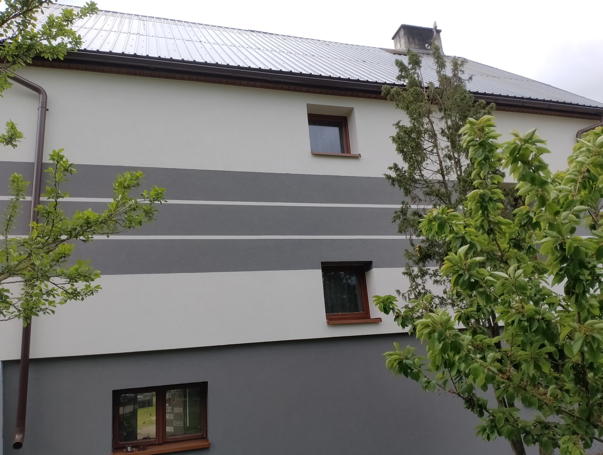 Elewacja domu z poziomymi pasami w odcieniach szarości i bieli, okna z brązowymi ramami, metalowy dach i zieleń drzew w kadrze.