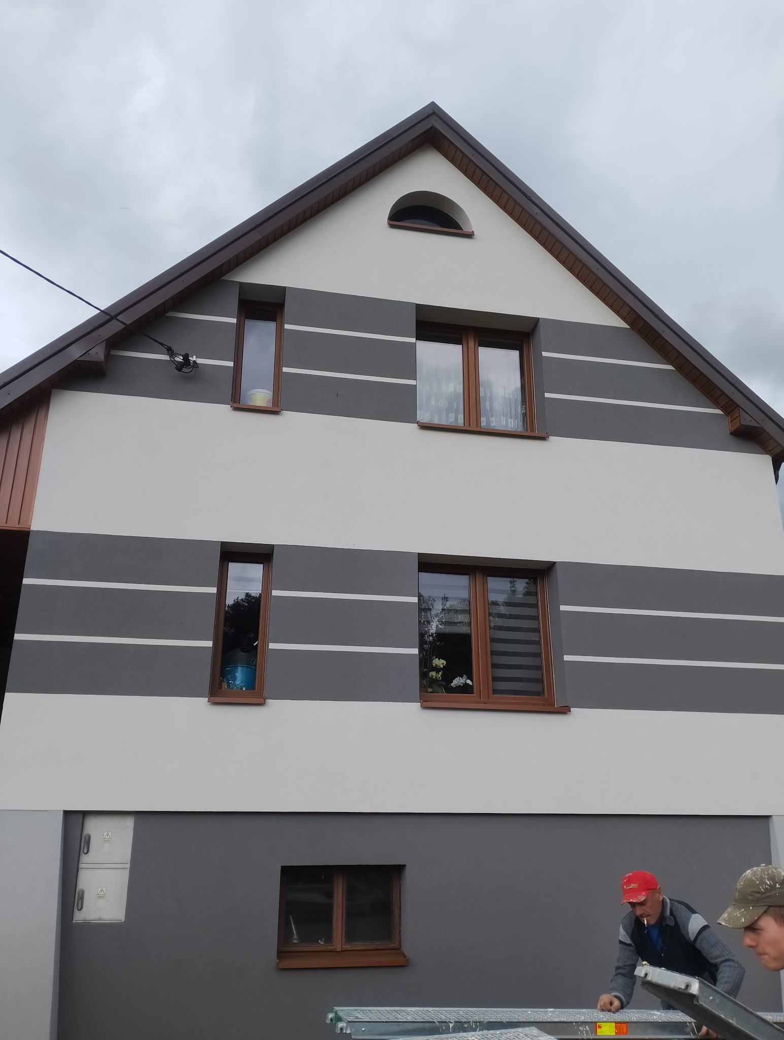 Dwukondygnacyjny dom z szarą i białą elewacją w poziome pasy. Widoczne brązowe okna, dwaj pracownicy na drabinie i pochmurne niebo.