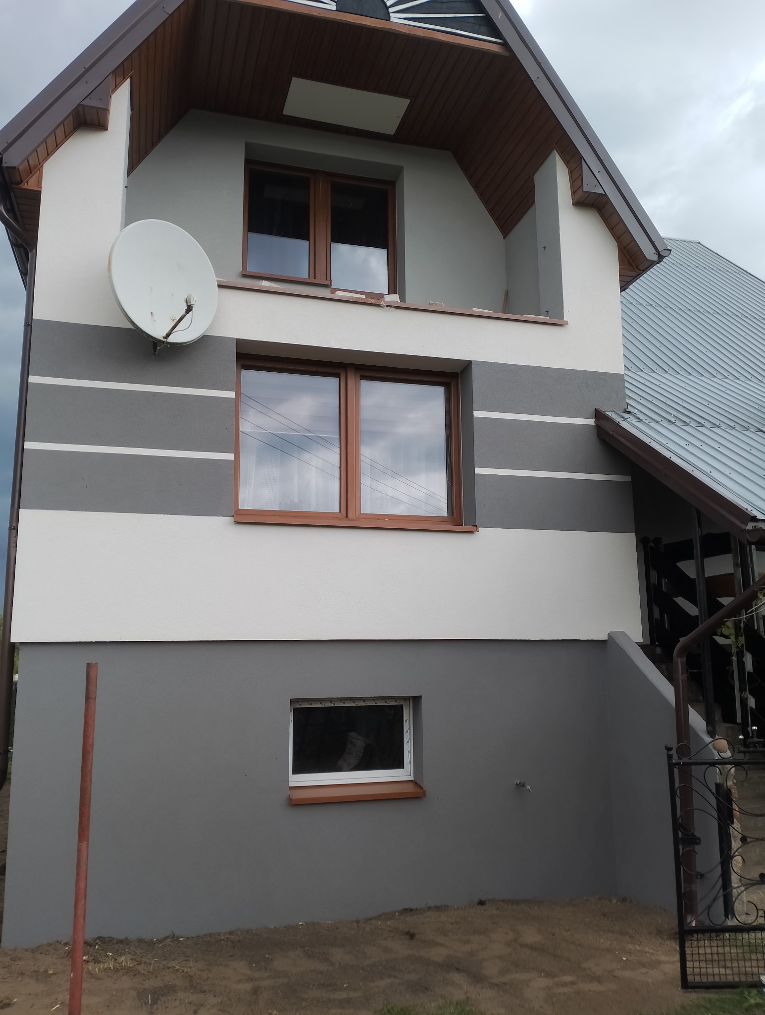 Dwukondygnacyjny dom z szarą elewacją w poziome pasy i brązowymi ramami okiennymi, antena satelitarna na ścianie. Widoczne elementy dachu i rynny.