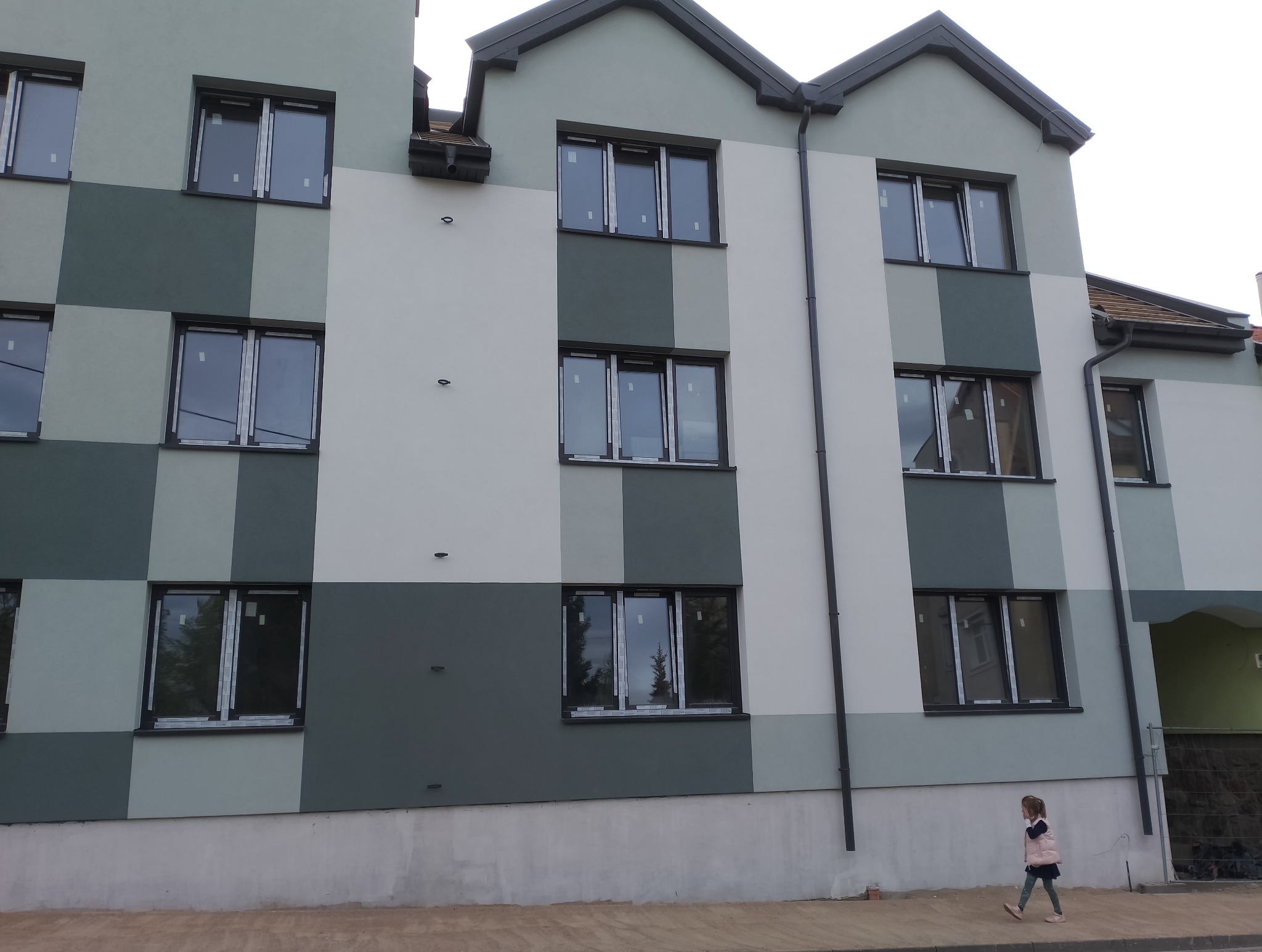 Nowoczesna elewacja budynku wielorodzinnego w odcieniach szarości i bieli, z widocznymi oknami i małą dziewczynką spacerującą wzdłuż chodnika.