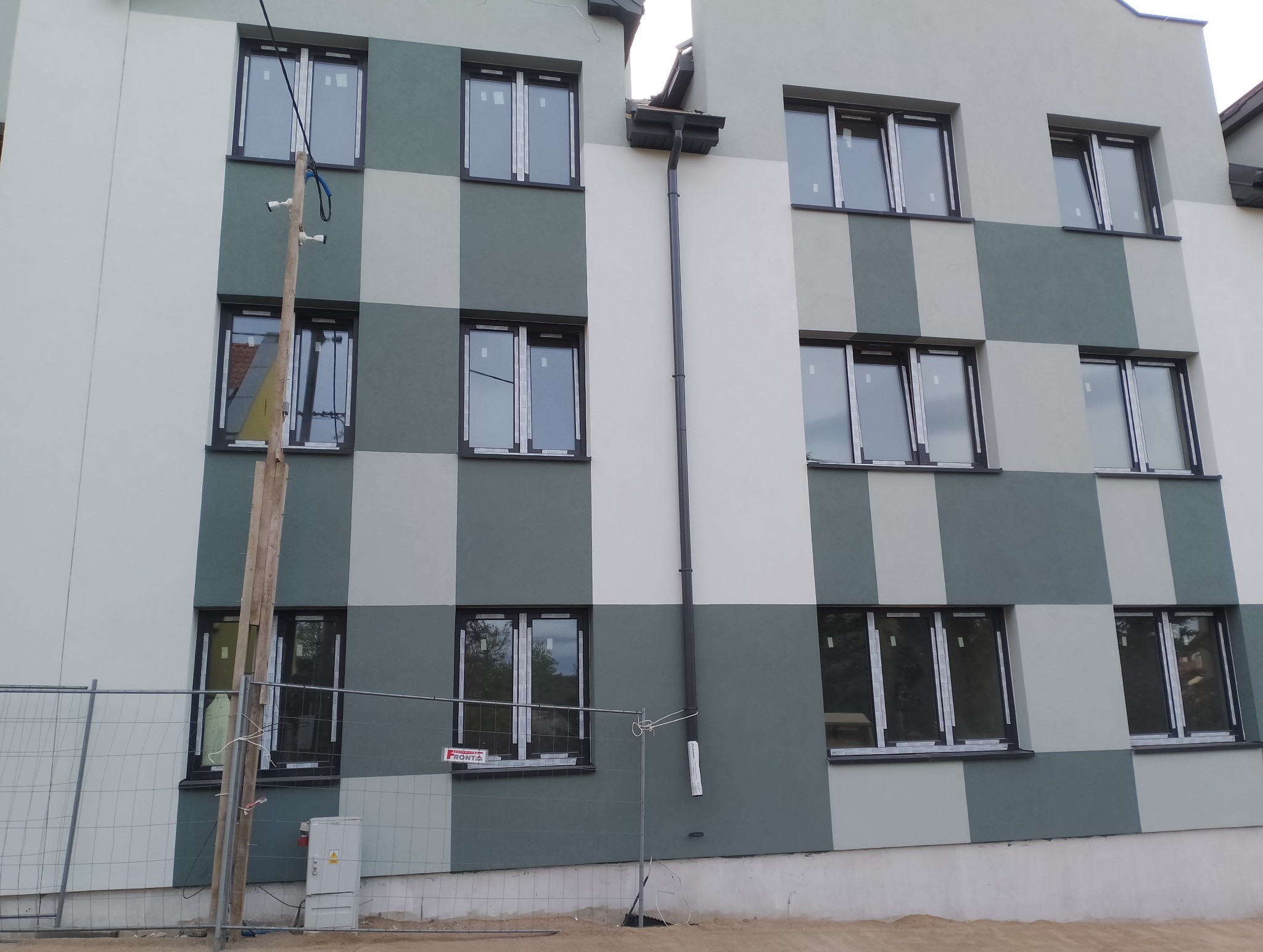 Elewacja budynku w szachownicę, świeżo po tynkowaniu, z nowymi oknami zabezpieczonymi folią. Widoczny fragment rusztowania i słup z kablami.