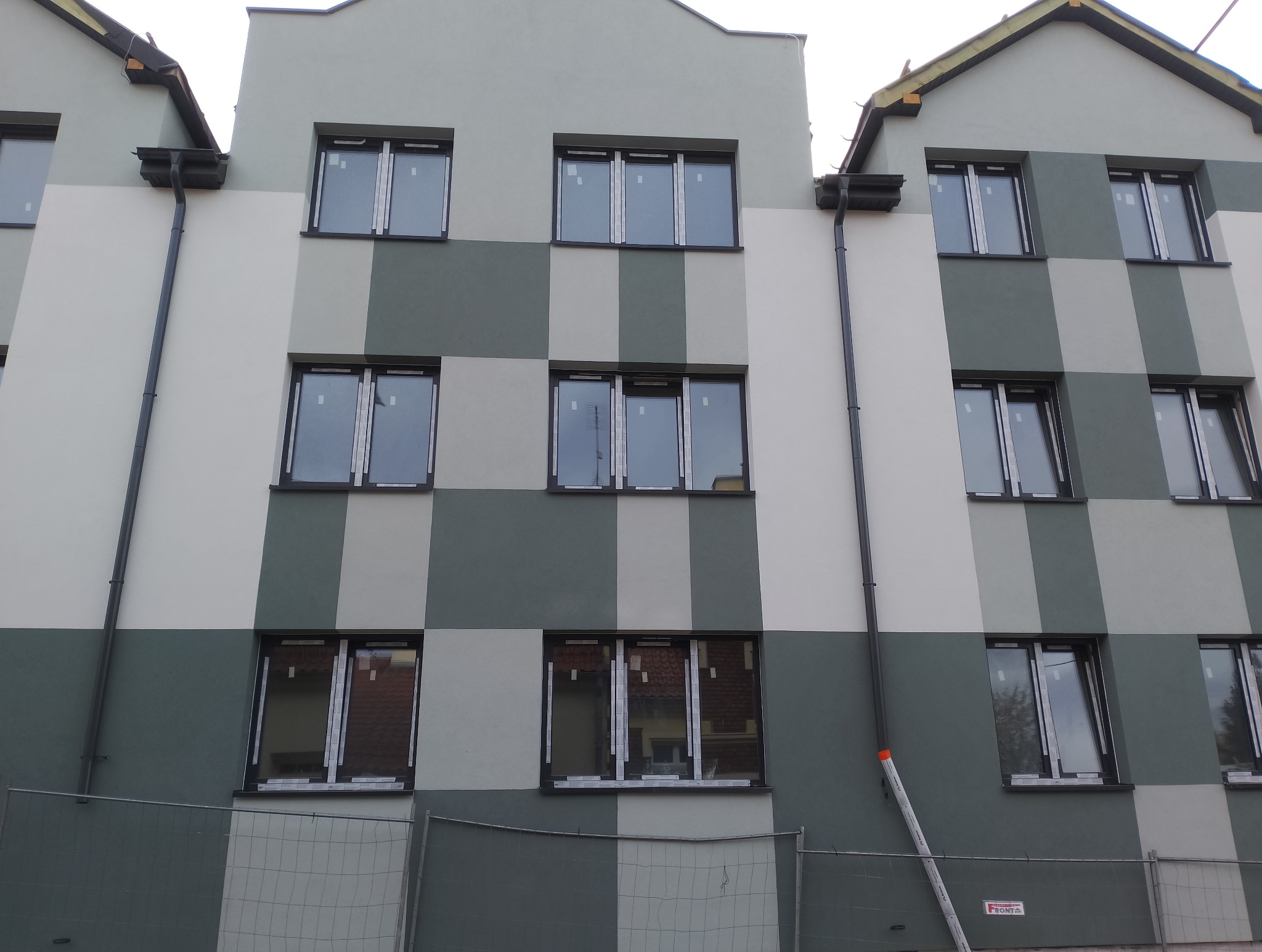 Wielopiętrowy budynek z elewacją w szachownicę, w odcieniach szarości. Widoczne okna z zabezpieczeniami i rynny. Całość ogrodzona siatką.