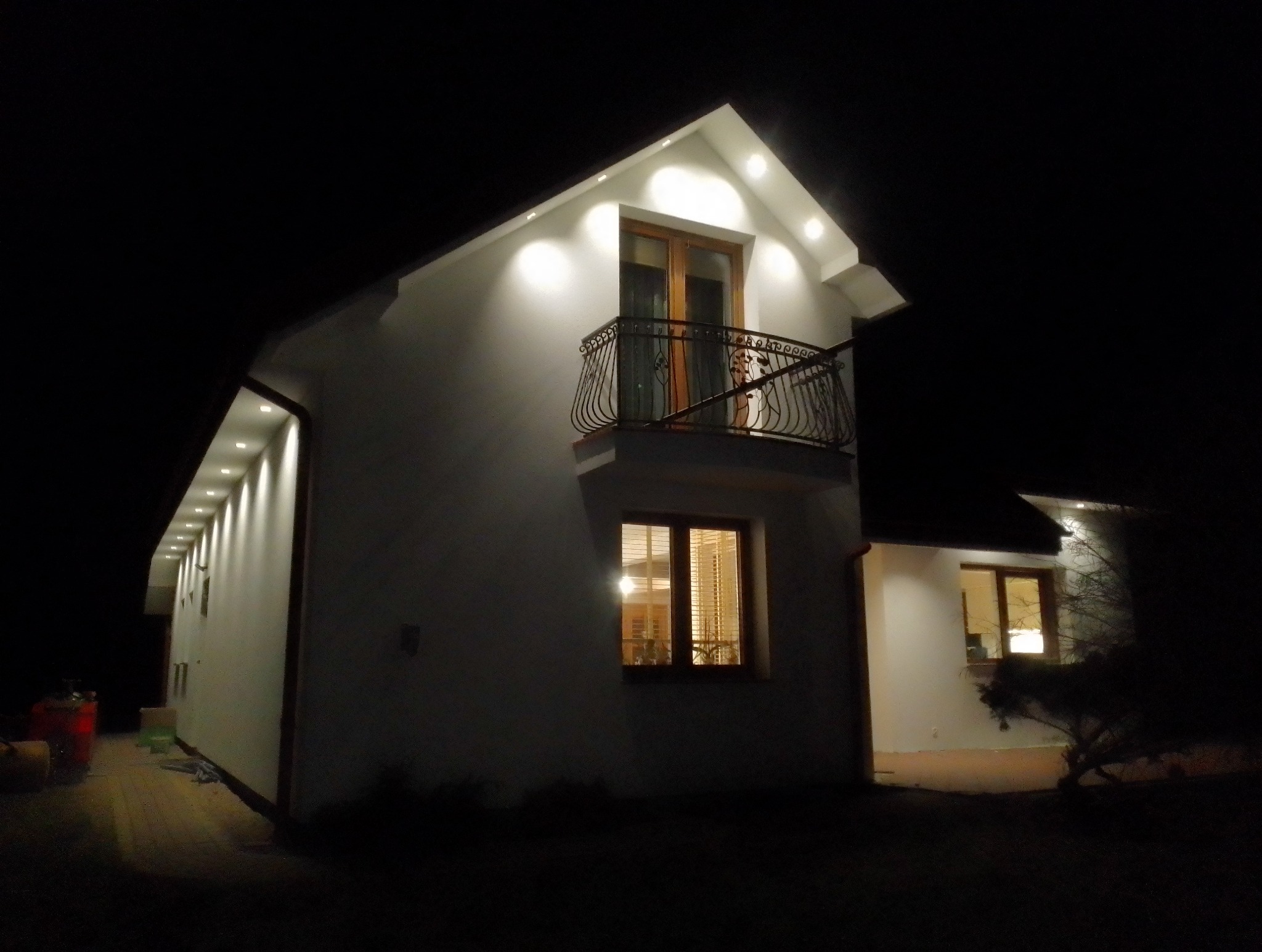 Nocne ujęcie oświetlonej elewacji domu z balkonem. Biała fasada z dekoracyjnym oświetleniem punktowym. Ciemne tło podkreśla detale architektoniczne i dodaje elegancji.