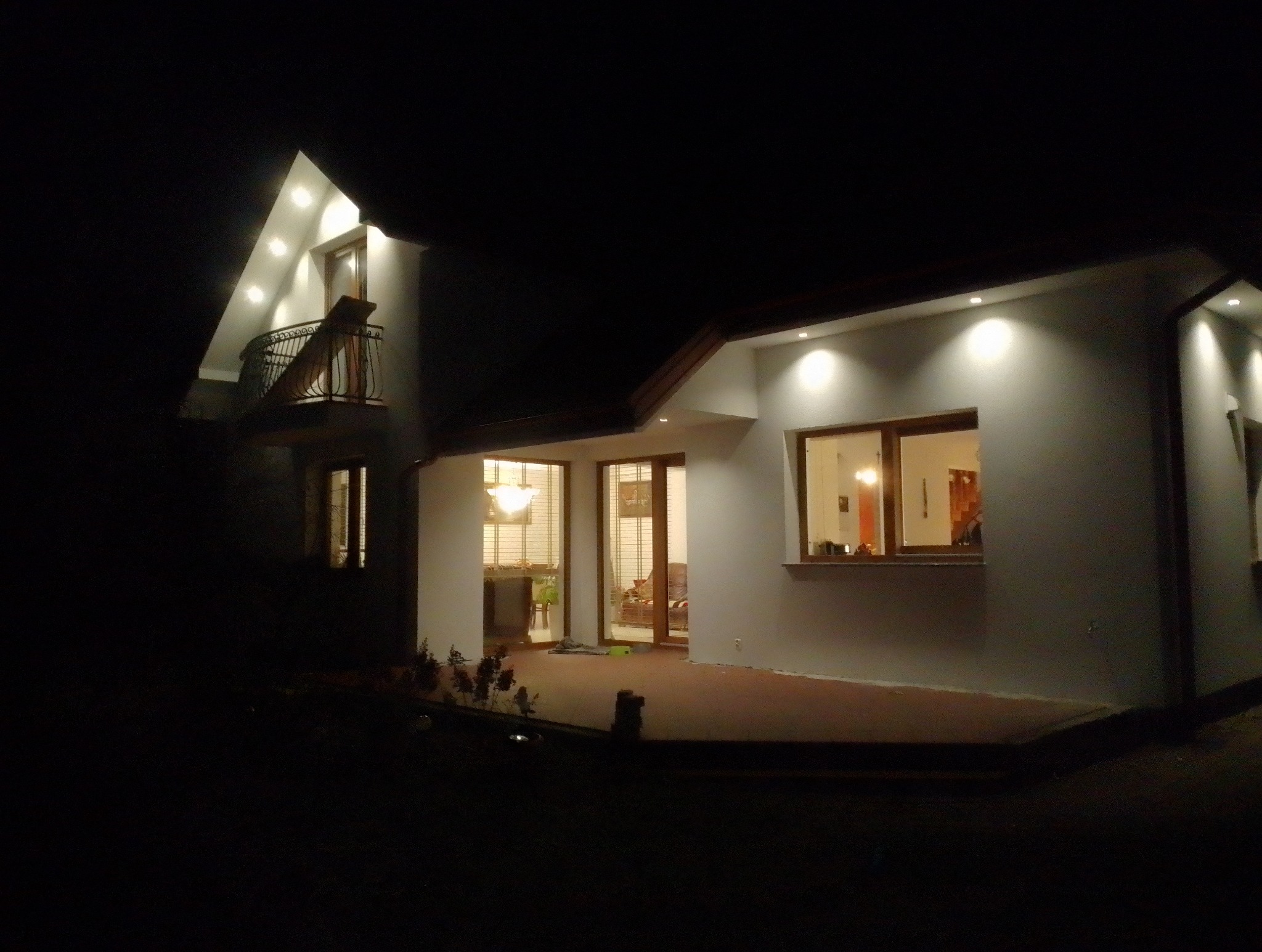 Oświetlony nocą dom jednorodzinny z balkonem i jasną elewacją. Widoczne okna i drzwi z drewnianą stolarką. Oświetlenie elewacyjne punktowe.