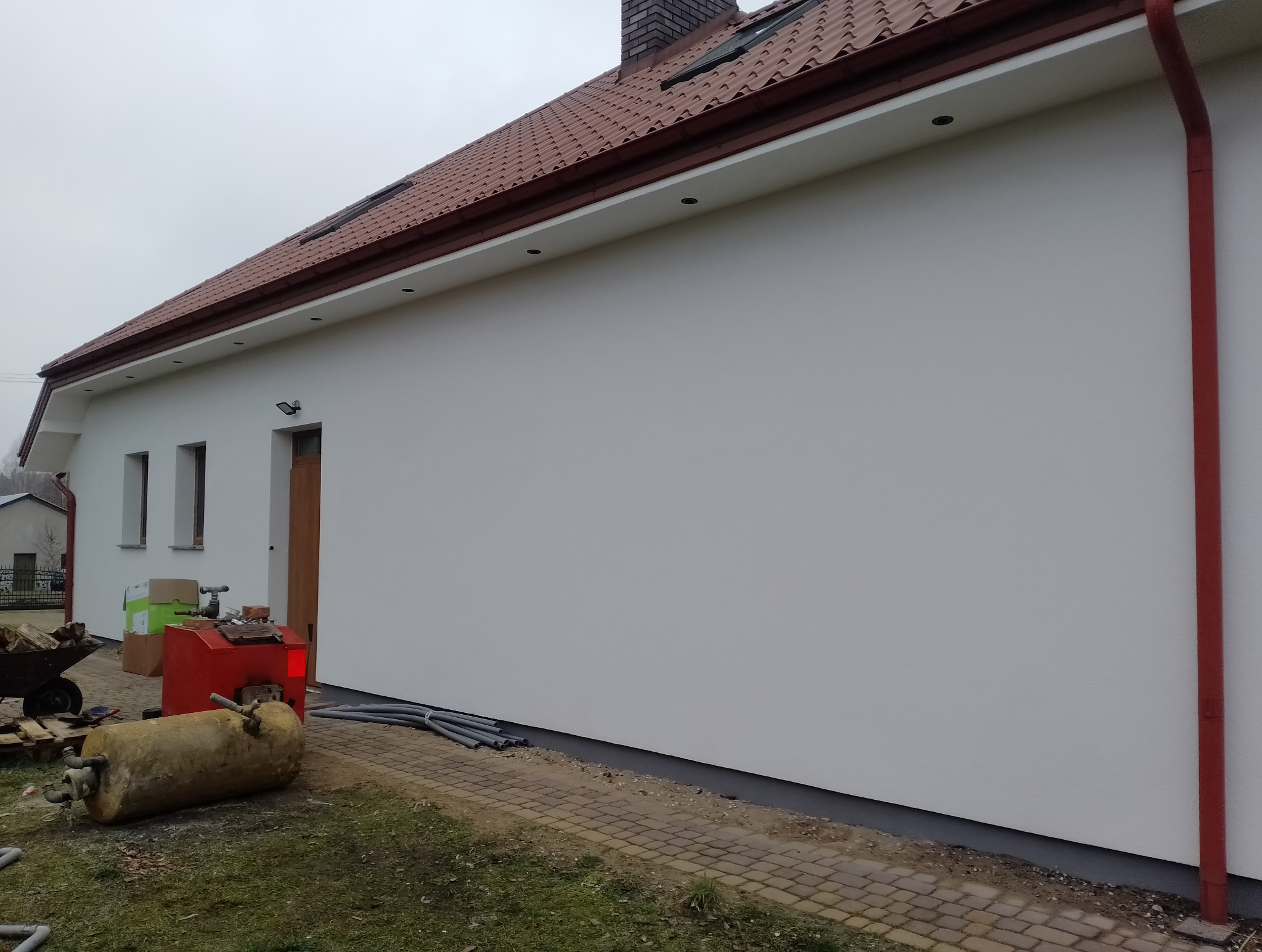 Biała, gładka elewacja domu jednorodzinnego z brązowym dachem i czerwoną rynną. Widoczne okna, drzwi wejściowe i elementy instalacji na zewnątrz budynku.