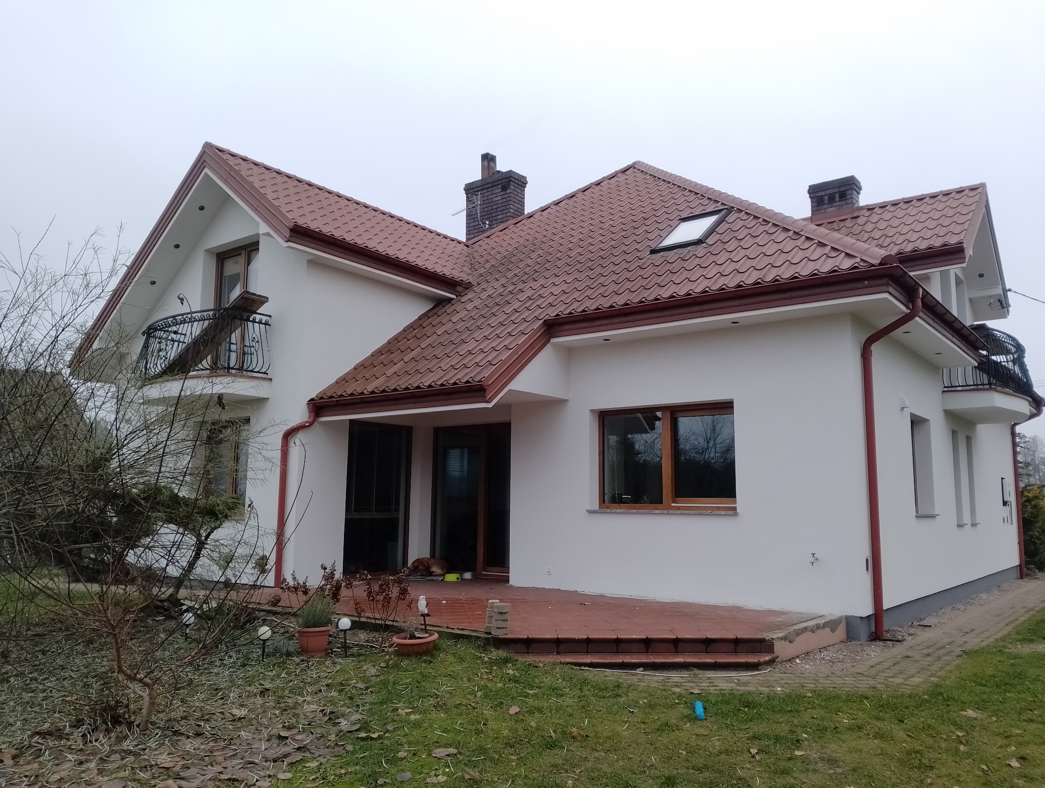 Biały dom jednorodzinny z czerwoną dachówką, widok z zewnątrz. Na tarasie doniczki i leżący pies. Elementy wykończeniowe w kolorze dachu.
