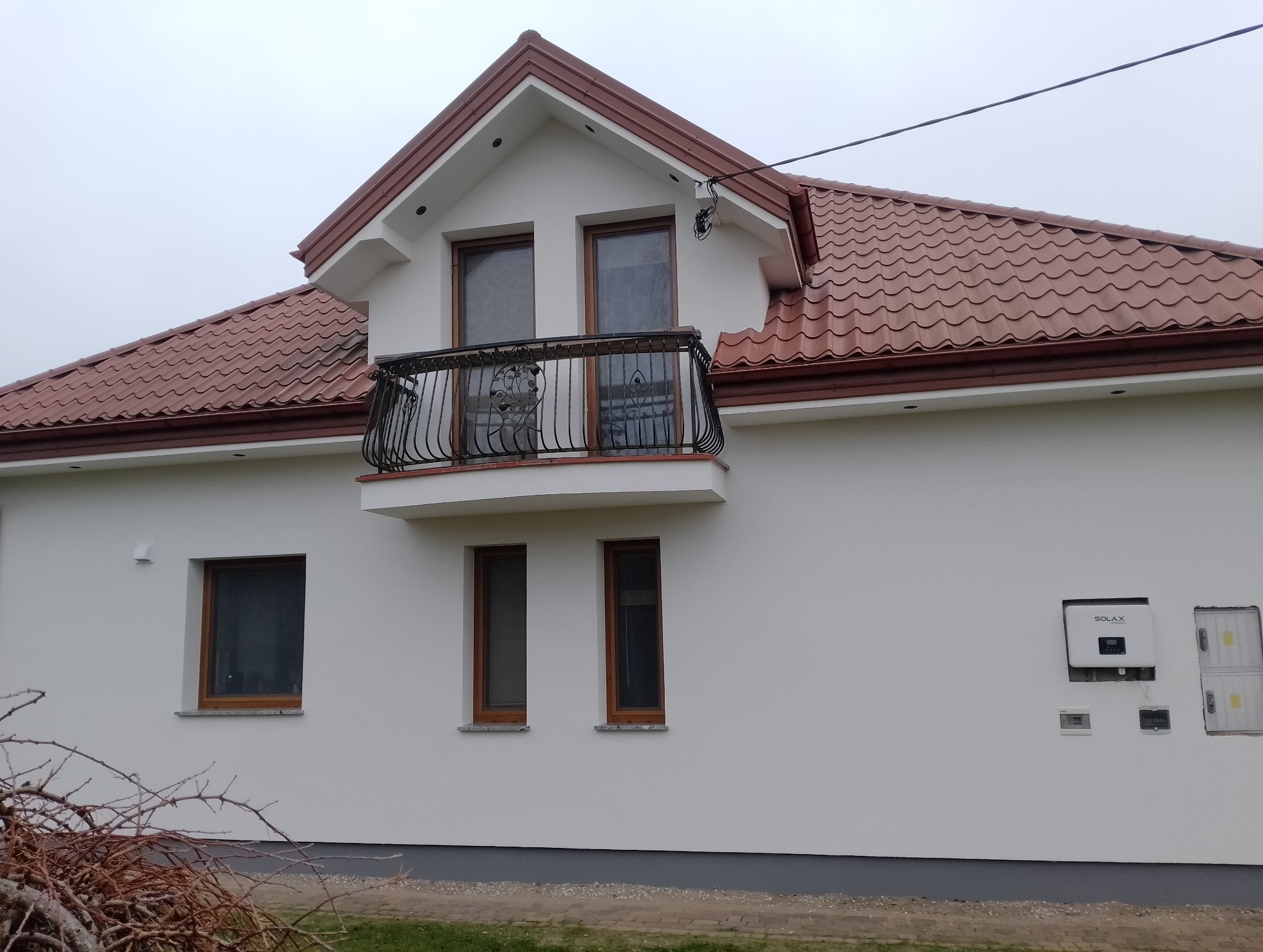 Biały dom z balkonem i czerwoną dachówką. Widoczny panel solarny na elewacji. Ujęcie z dołu, pochmurne niebo w tle. Elementy dekoracyjne balkonu.