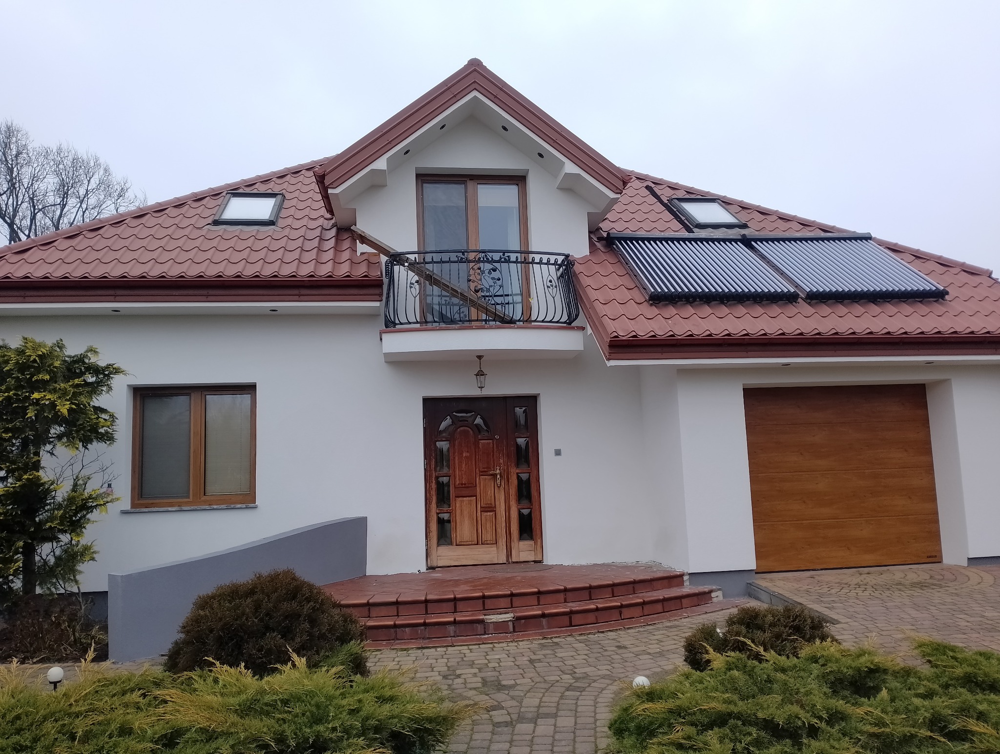 Biały dom jednorodzinny z czerwoną dachówką, panelami solarnymi i drewnianymi elementami wykończeniowymi. Widoczny balkon z ozdobną balustradą i zadbany ogród.