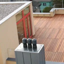 Nowoczesny dach płaski wykończony drewnianymi panelami, z widocznymi elementami obróbek blacharskich i kominem wentylacyjnym, pokryty żwirem na części powierzchni.