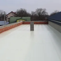 Przygotowana powierzchnia dachu pokryta białym materiałem izolacyjnym, obrzeże z cegieł i szara blacha dachowa w tle krajobraz wiejski.