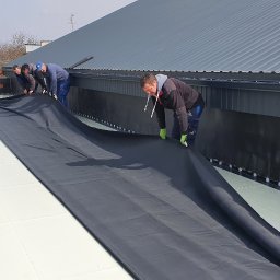 TOMDACH TOMASZ WŁODARCZYK - Ekipa dekarzy rozkłada czarną membranę hydroizolacyjną na płaskim dachu z widocznymi elementami mocującymi, w tle blaszany dach i budynki.