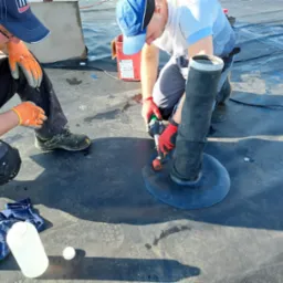 Dwóch dekarzy pracuje na płaskim dachu, uszczelniając przejście rury wentylacyjnej za pomocą zgrzewarki dekarskiej i membrany bitumicznej.