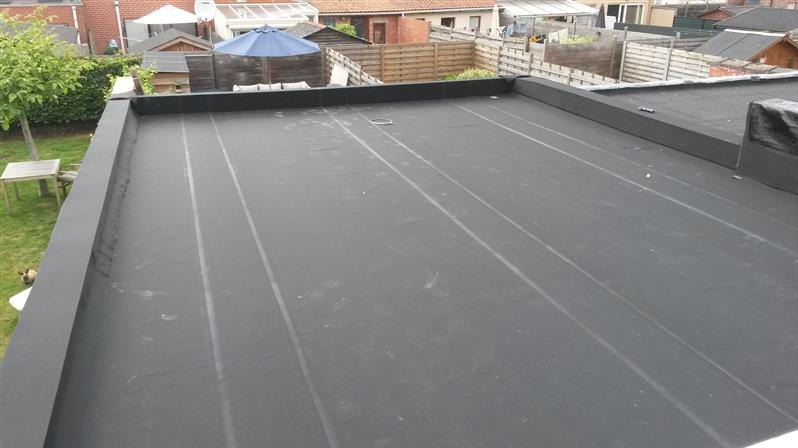 Nowe, płaskie pokrycie dachu z papy termozgrzewalnej, widoczne obróbki blacharskie na krawędziach, w tle zabudowa jednorodzinna i ogród z parasolem.
